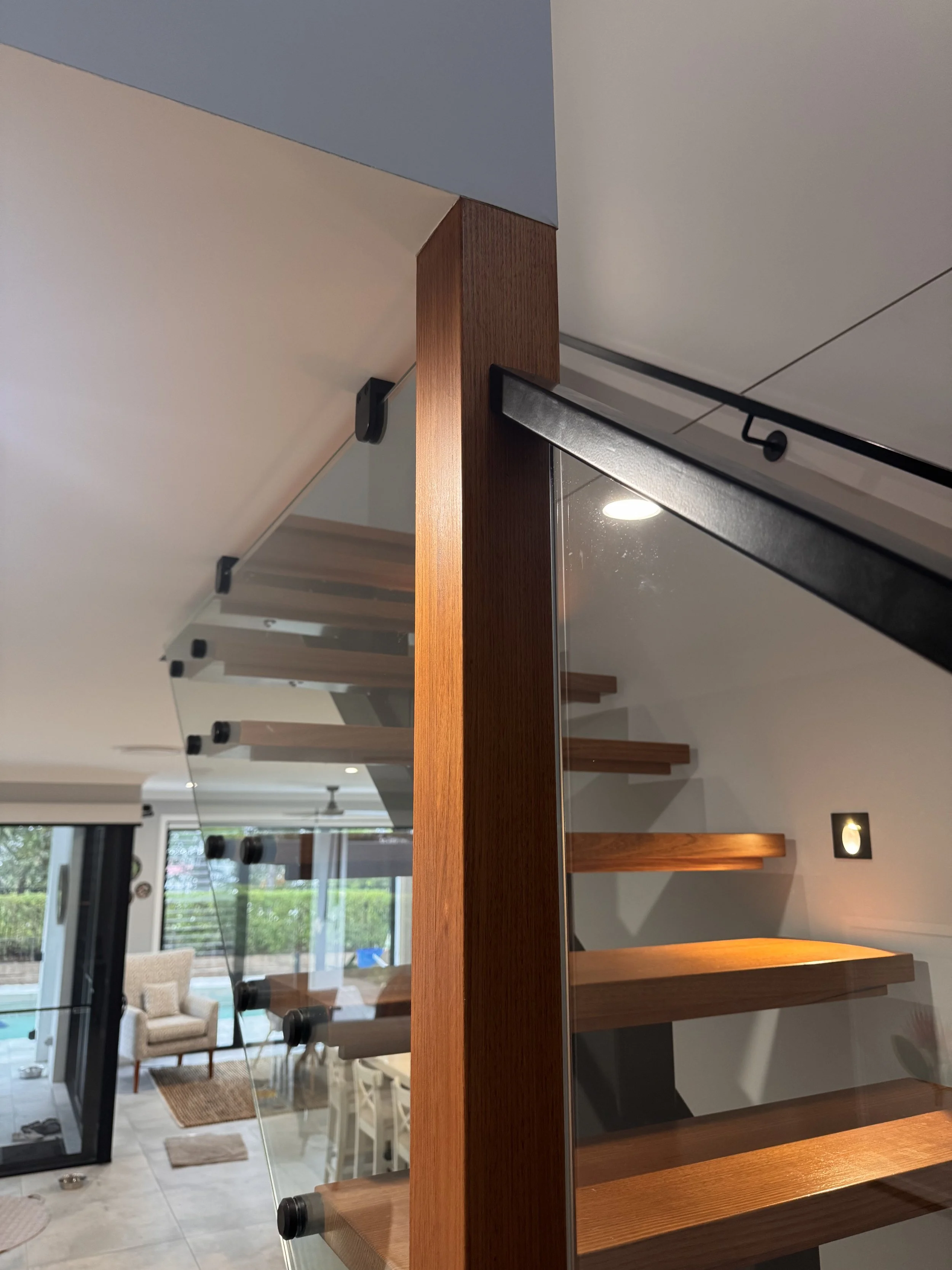 View of an indoor living space with a focus on a wooden and glass divider or shelving unit, with a glimpse of a living room area in the background including chairs, a table, and large windows.