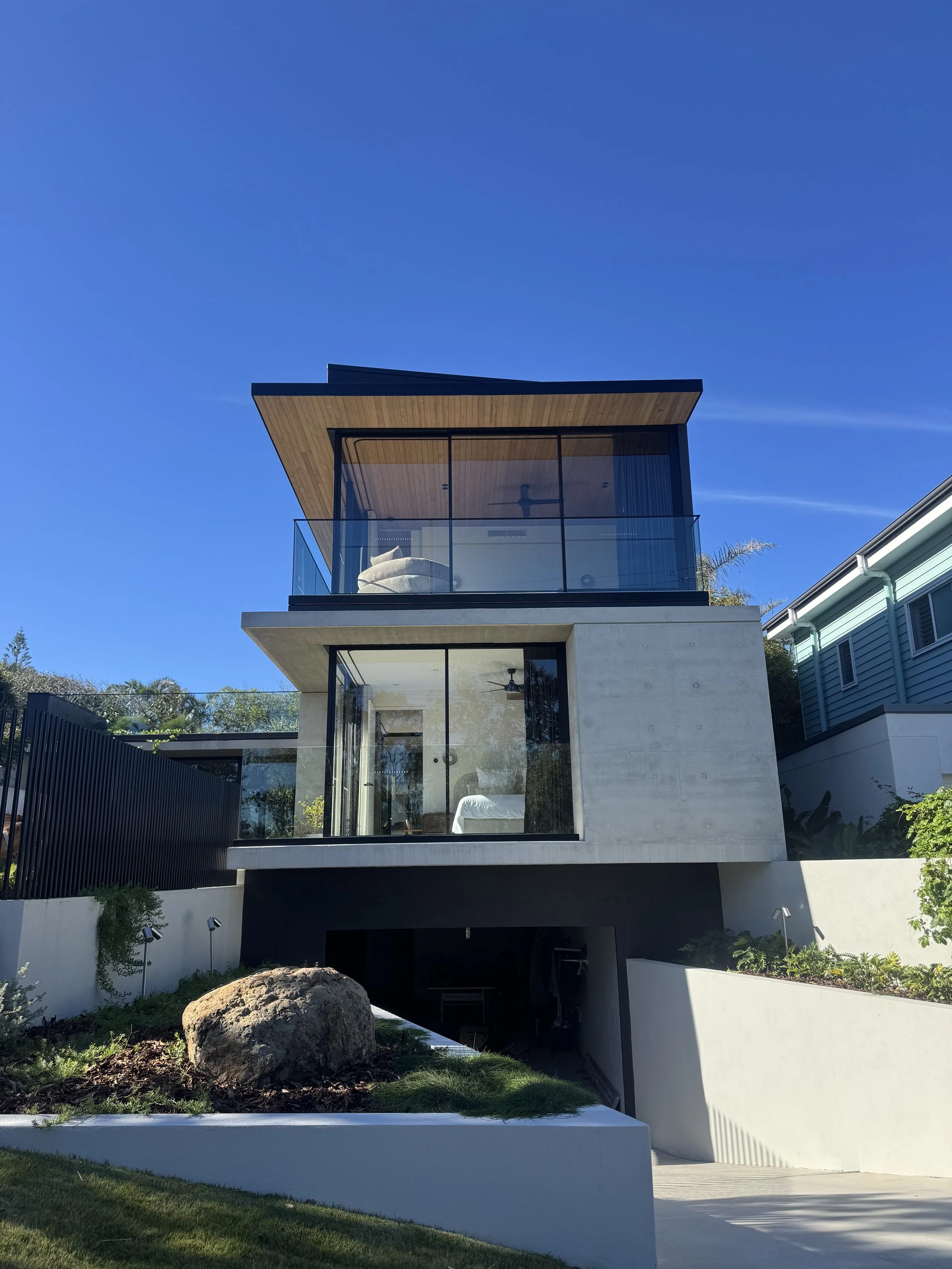 Modern multi-story house with large glass windows, concrete walls, and a wooden roof, surrounded by a landscaped yard with grass, a rock, and plants. The house features a balcony with a glass railing and a ceiling fan visible through the windows.