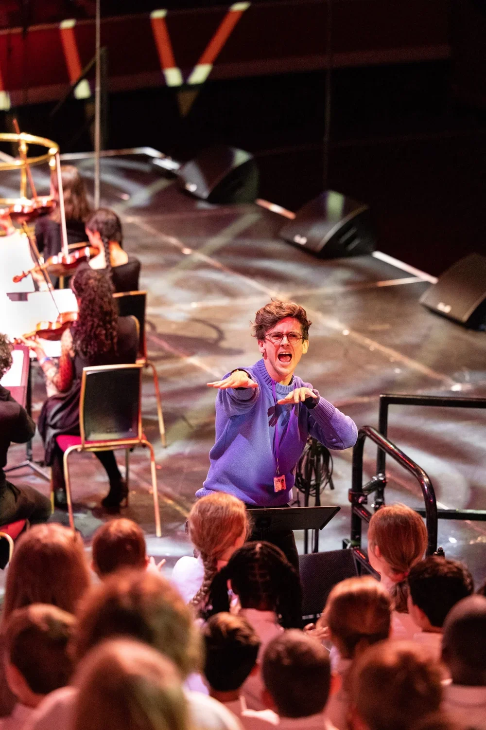 A man wearing glasses and a purple sweater is gesturing while speaking to an audience in a theater or auditorium. Behind him, there are several seated people, some with braided hair, and an orchestra with musicians and musical stands.