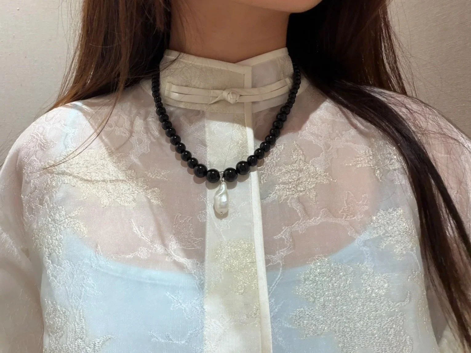 A woman wearing a cream-colored traditional blouse with a high collar and intricate floral patterns, adorned with a black beaded necklace with a large pearl pendant.