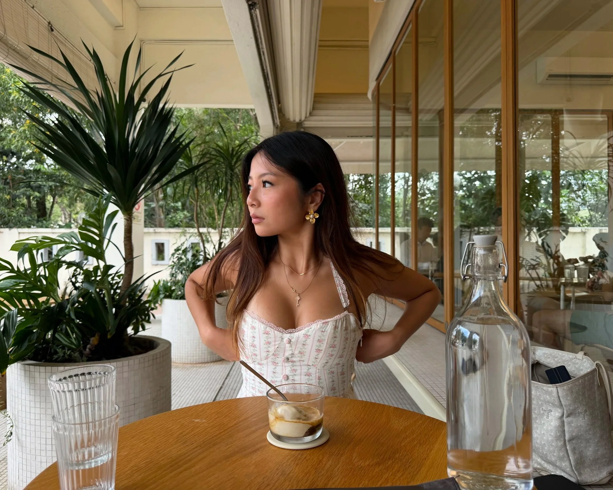 A woman with long brown hair wearing a white, floral-patterned dress and earrings sits at a wooden table with a dessert in front of her. The table is set with clear glasses and a glass bottle. Large green plants and a glass window with wooden frames are in the background, suggesting a cozy cafe or restaurant setting.
