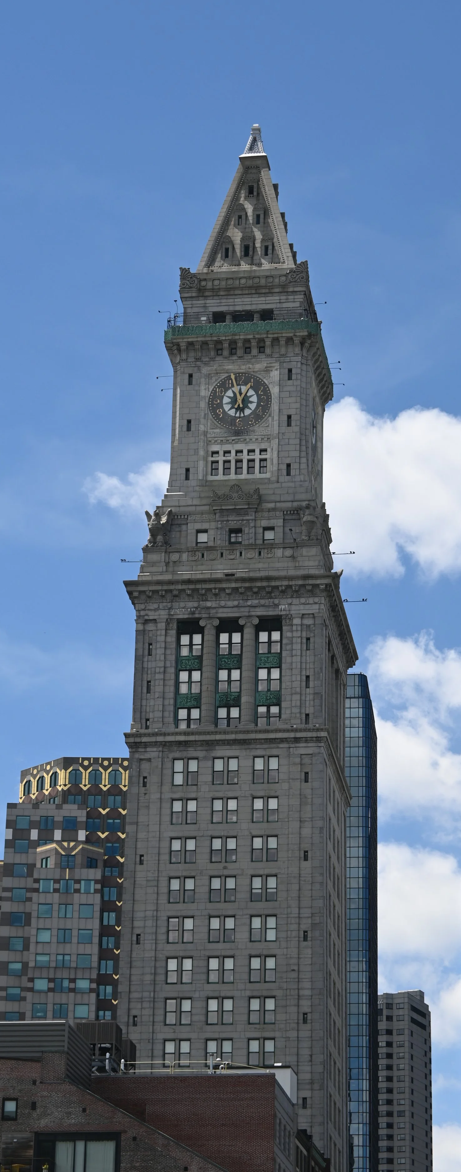 Tall clock tower with a spire in a cityscape, surrounded by modern buildings, against a blue sky with some clouds.