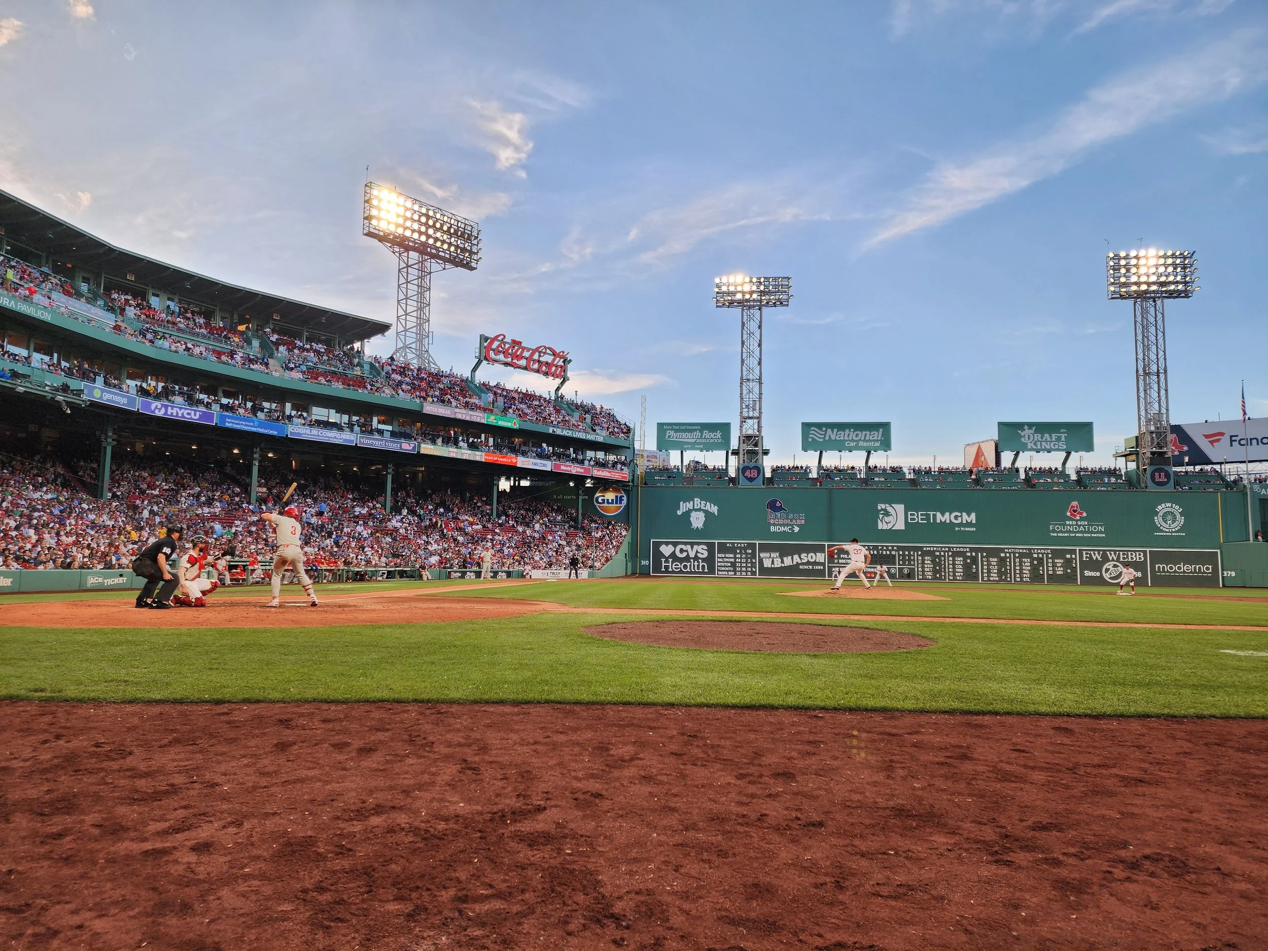 A baseball game at Fenway Park with players on the field and spectators in the stands, with the sky partly cloudy and stadium lights on.