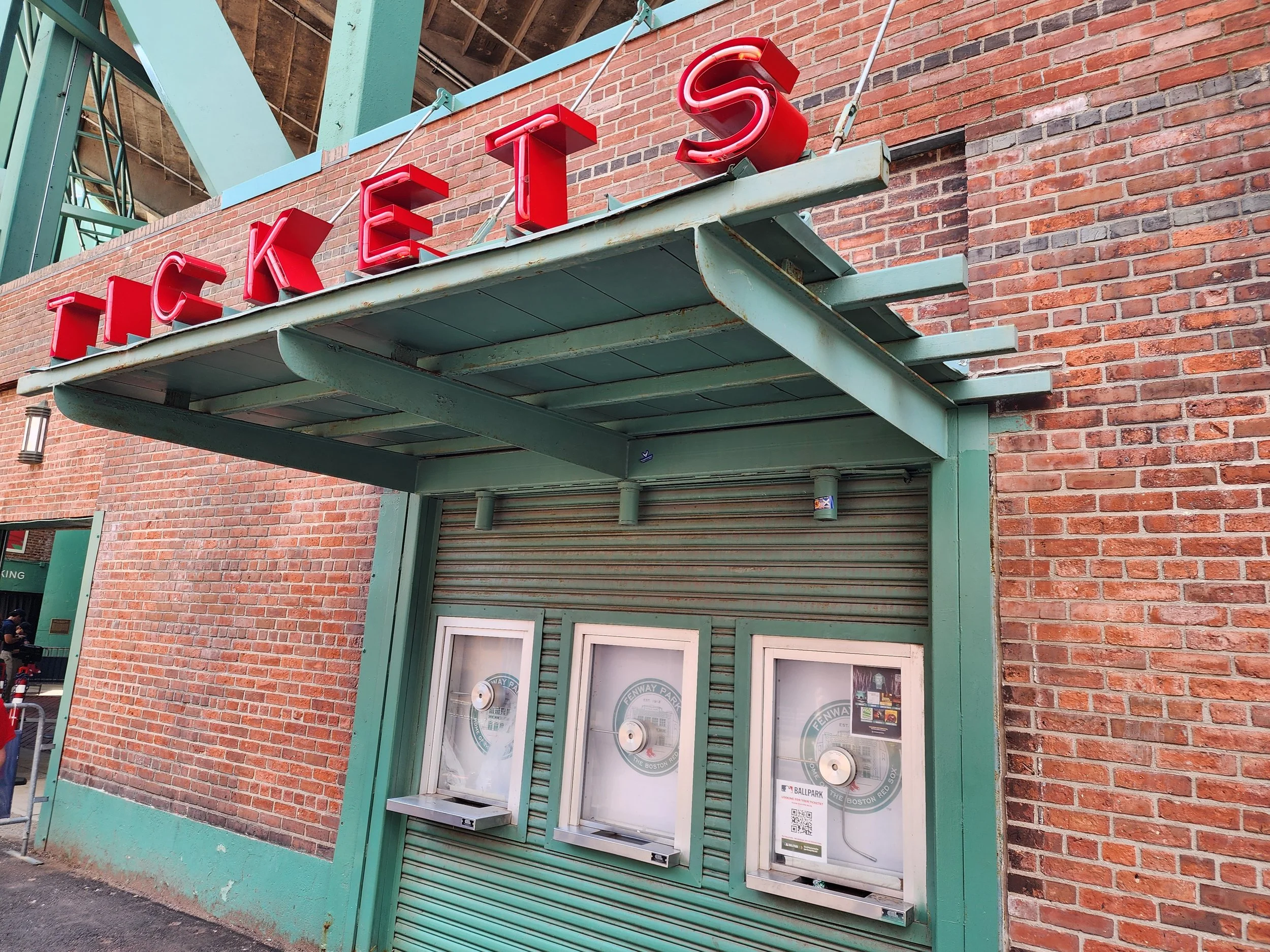 A closed storefront with a green metal awning and a brick wall background, featuring a red neon sign spelling 'TEA', with cafeteria-style ticket dispensers below.