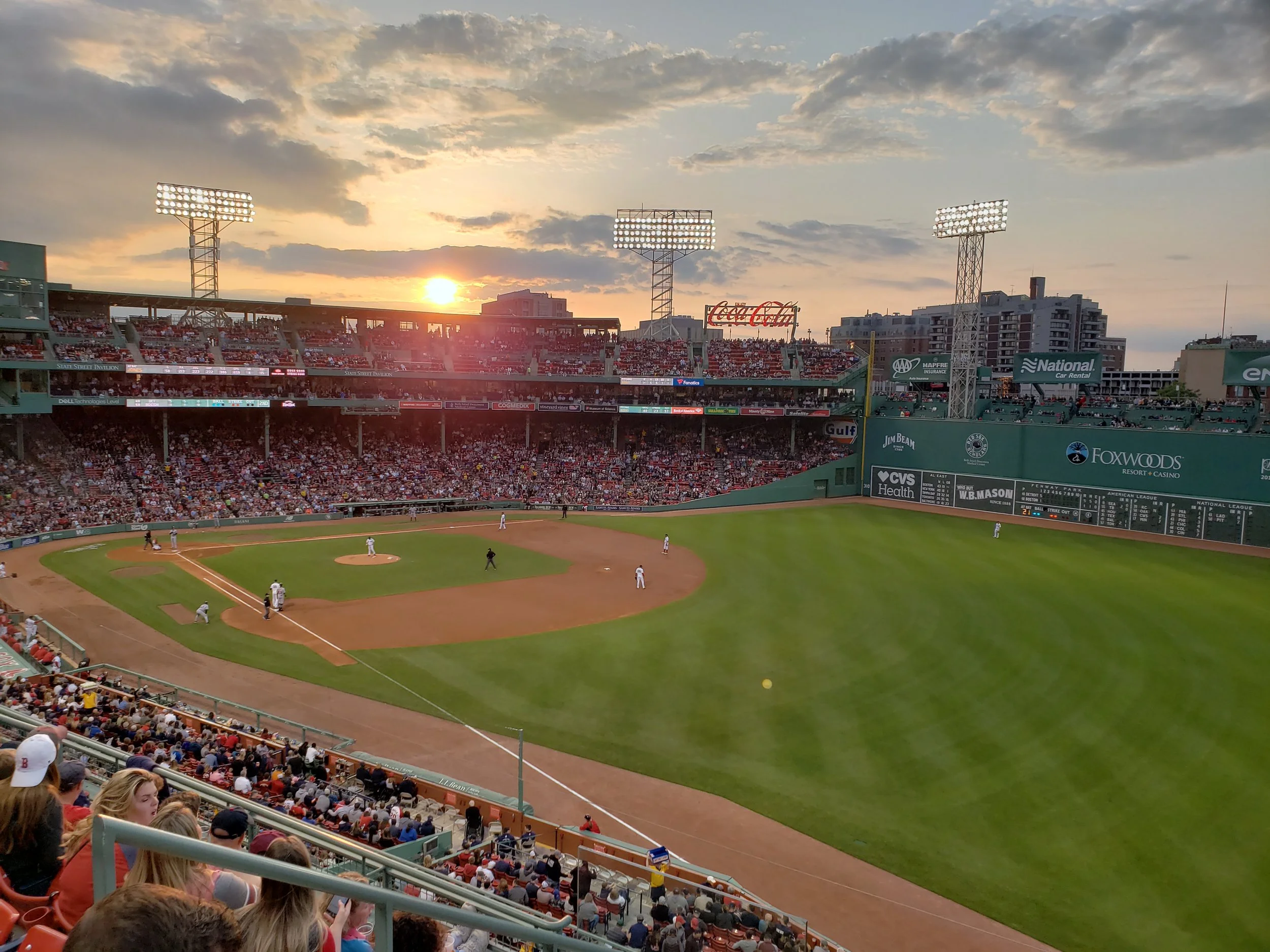 A baseball game at a stadium during sunset with fans in the stands and players on the field.