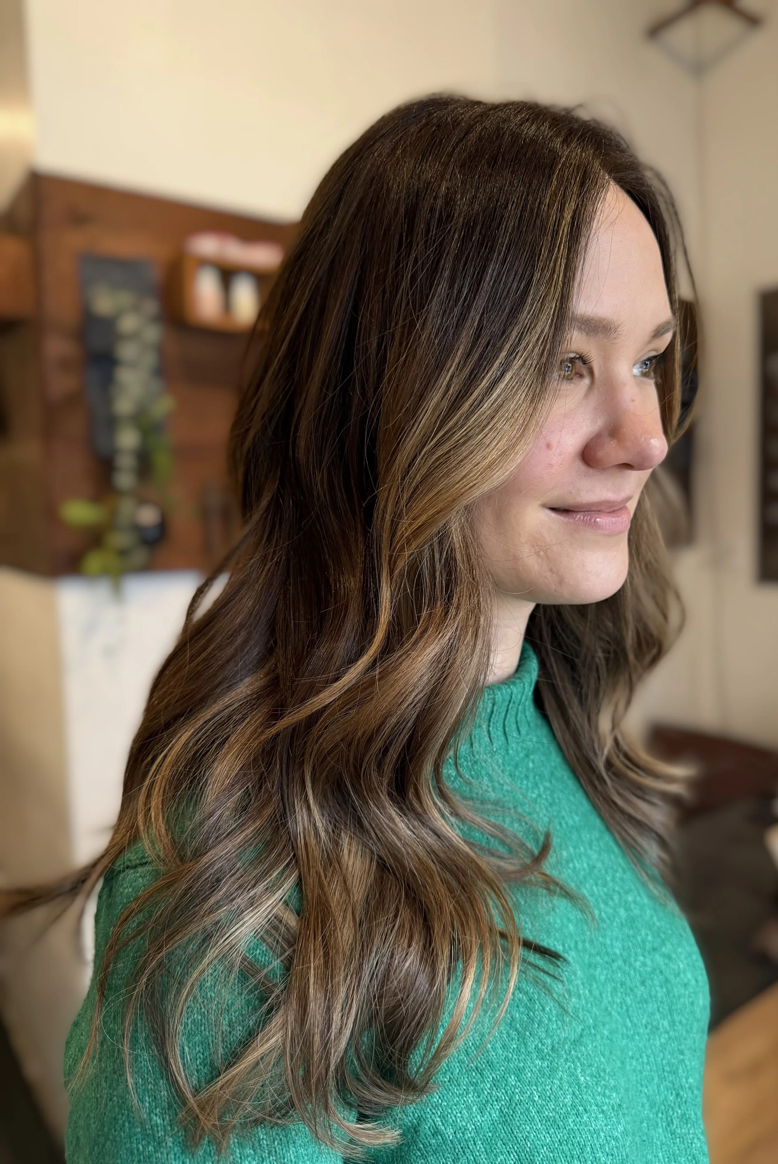 Close-up of a woman with long, wavy brown hair with blonde highlights, wearing a green sweater, smiling slightly, in a cozy indoor setting.