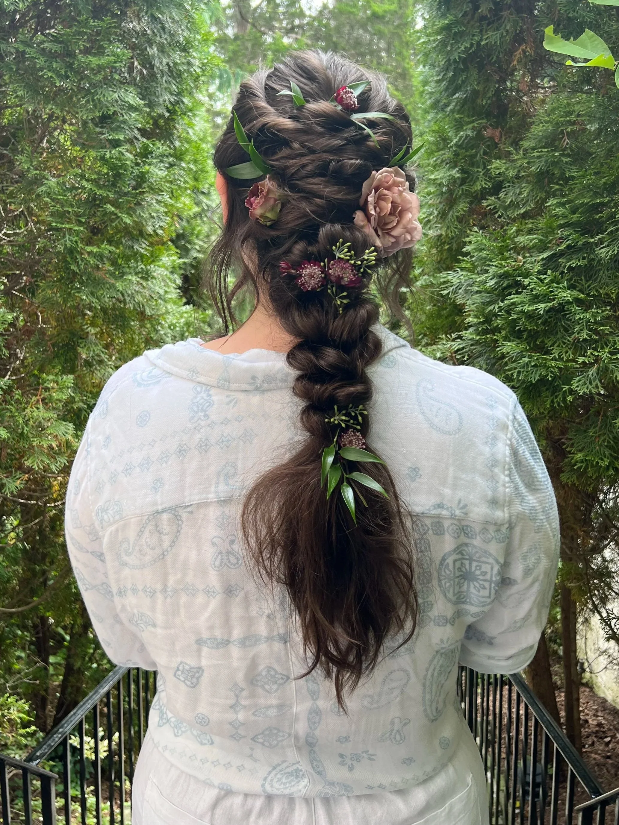 Back view of a person with long dark hair styled in a braid decorated with pink and burgundy flowers and green leaves, wearing a white pattern-embossed shirt, standing outdoors near green foliage.
