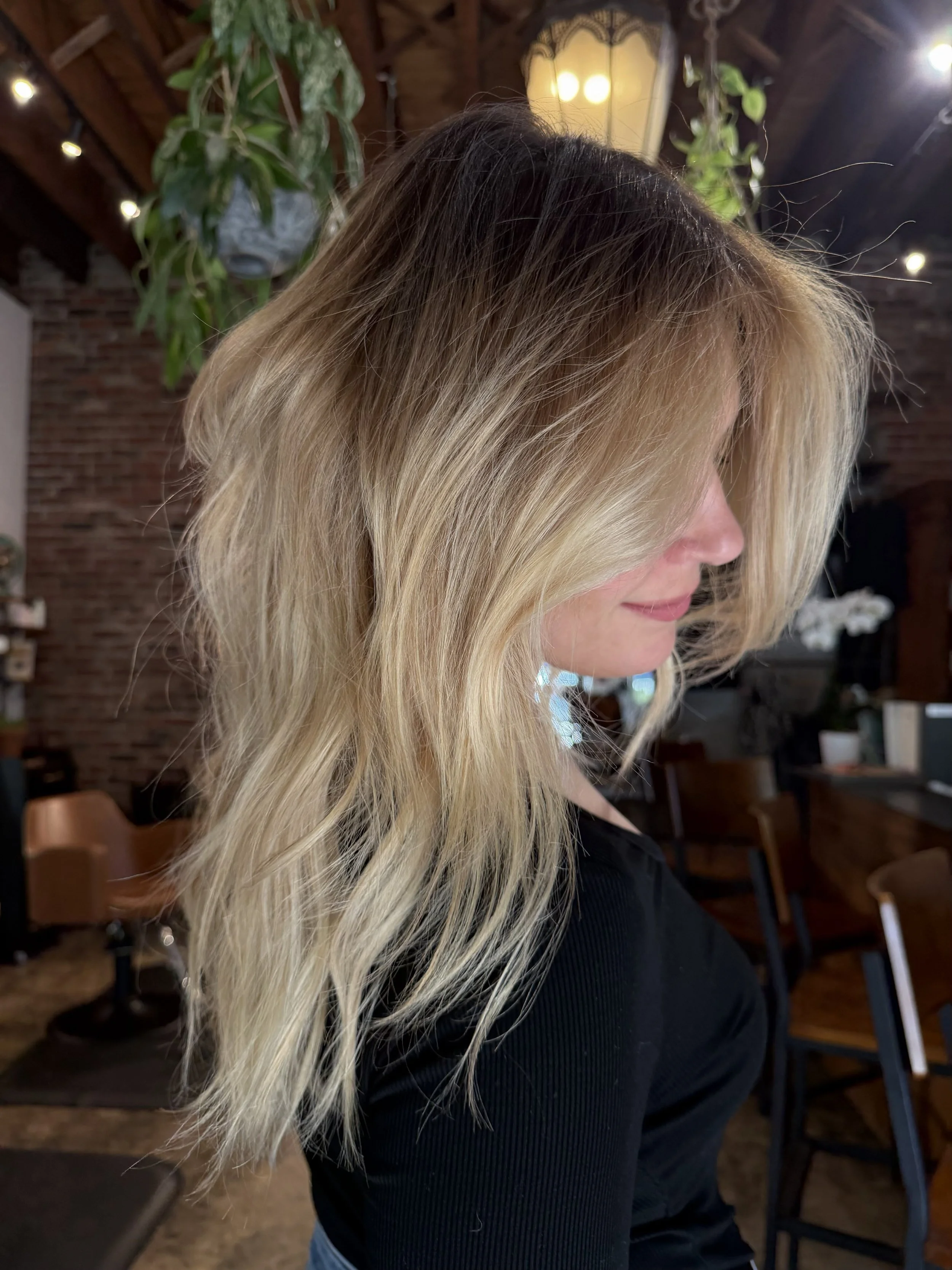 Side profile of a woman with long blonde hair, standing indoors with a brick wall and plants in the background.