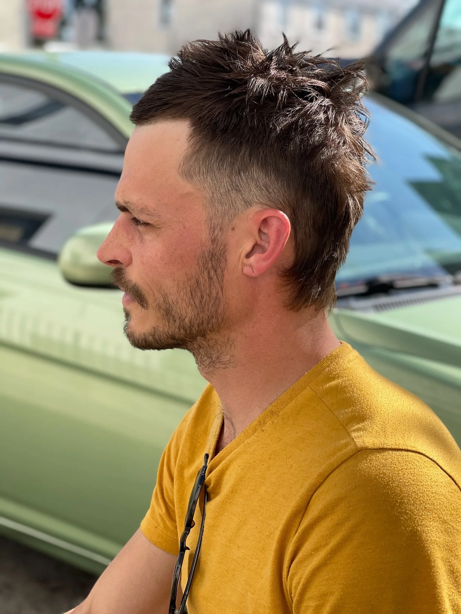 A young man with a short, textured haircut and facial hair, wearing a mustard yellow t-shirt, standing outdoors with a light green car and buildings in the background.