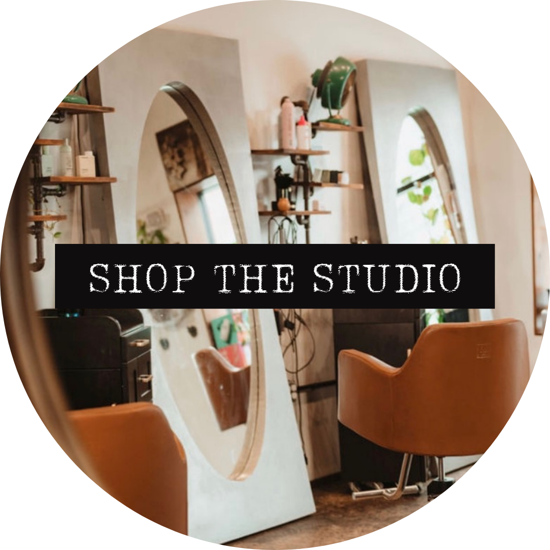 Interior view of a hair salon or barber shop with a large oval mirror, two brown chairs, shelves with hair products, and plants, with the text 'Shop the Studio' overlayed.