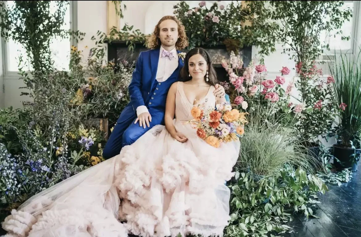 A couple dressed in wedding attire poses in front of a lush floral arrangement with pink roses and greenery, with the woman holding a bouquet of colorful flowers.
