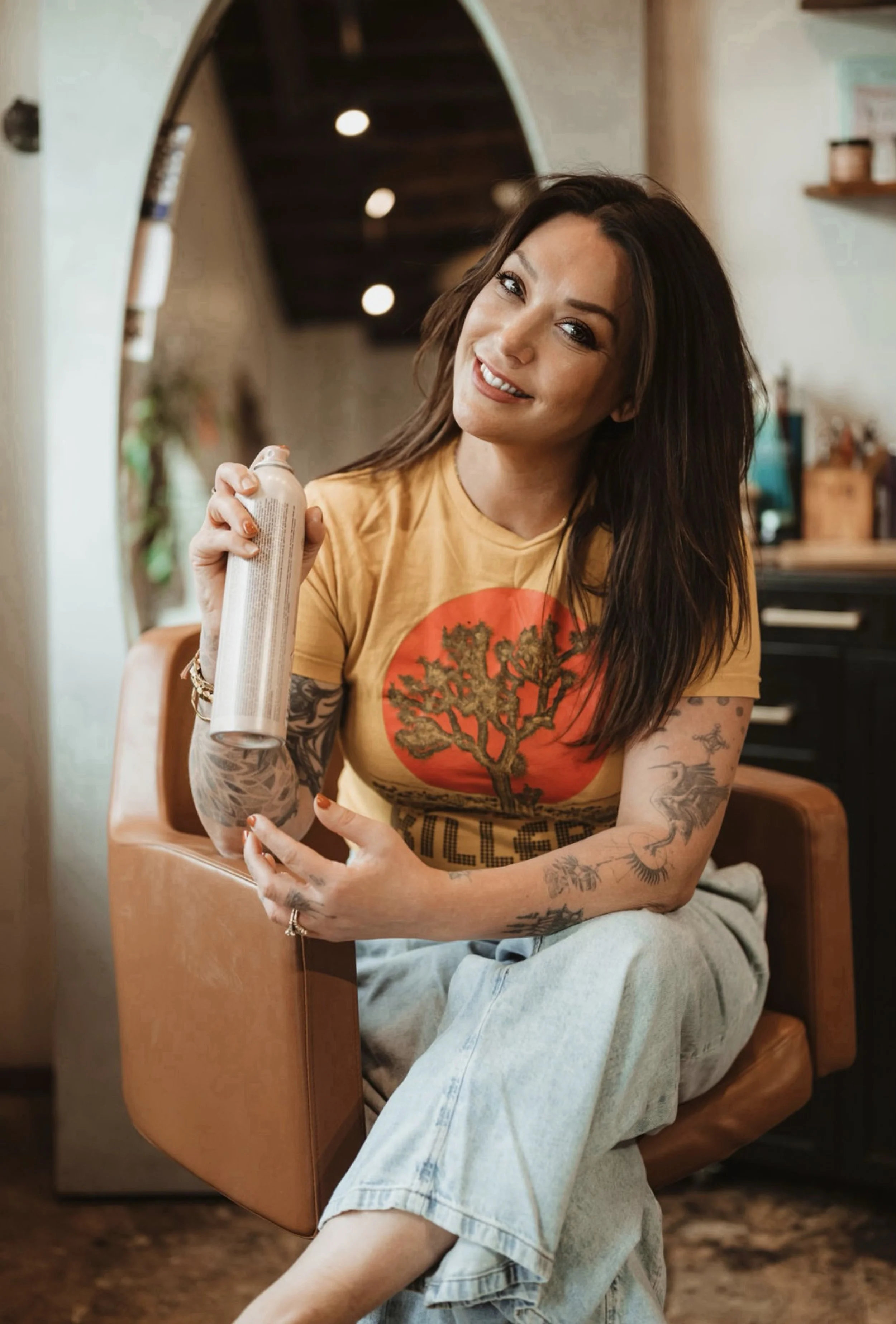 A woman with tattoos on her arms and long dark hair sitting on a brown leather chair, holding a can of spray in her right hand, smiling at the camera, inside a cozy room with warm lighting.
