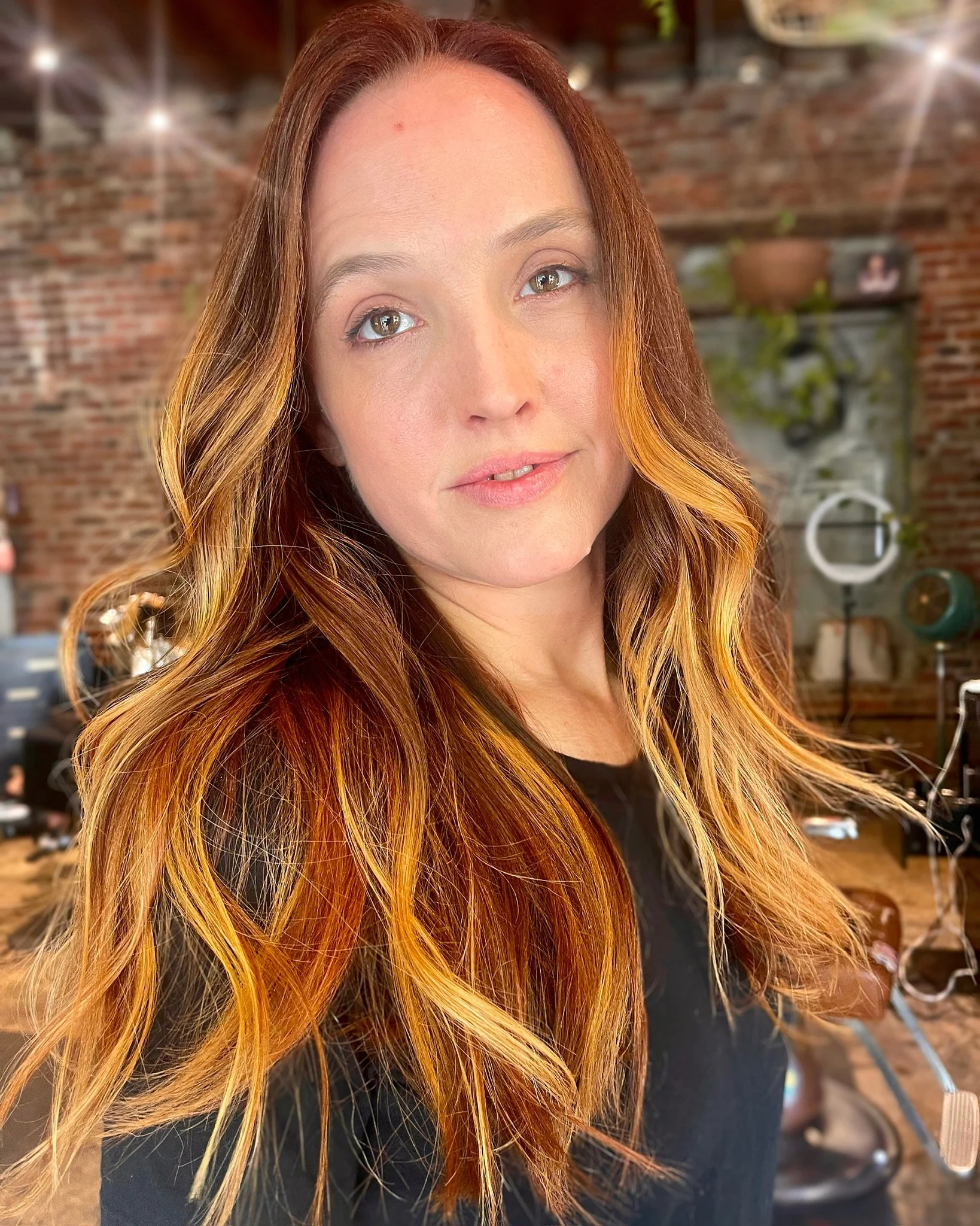 A woman with wavy, reddish-brown hair and light skin poses in a salon or studio with brick walls and art decor in the background.