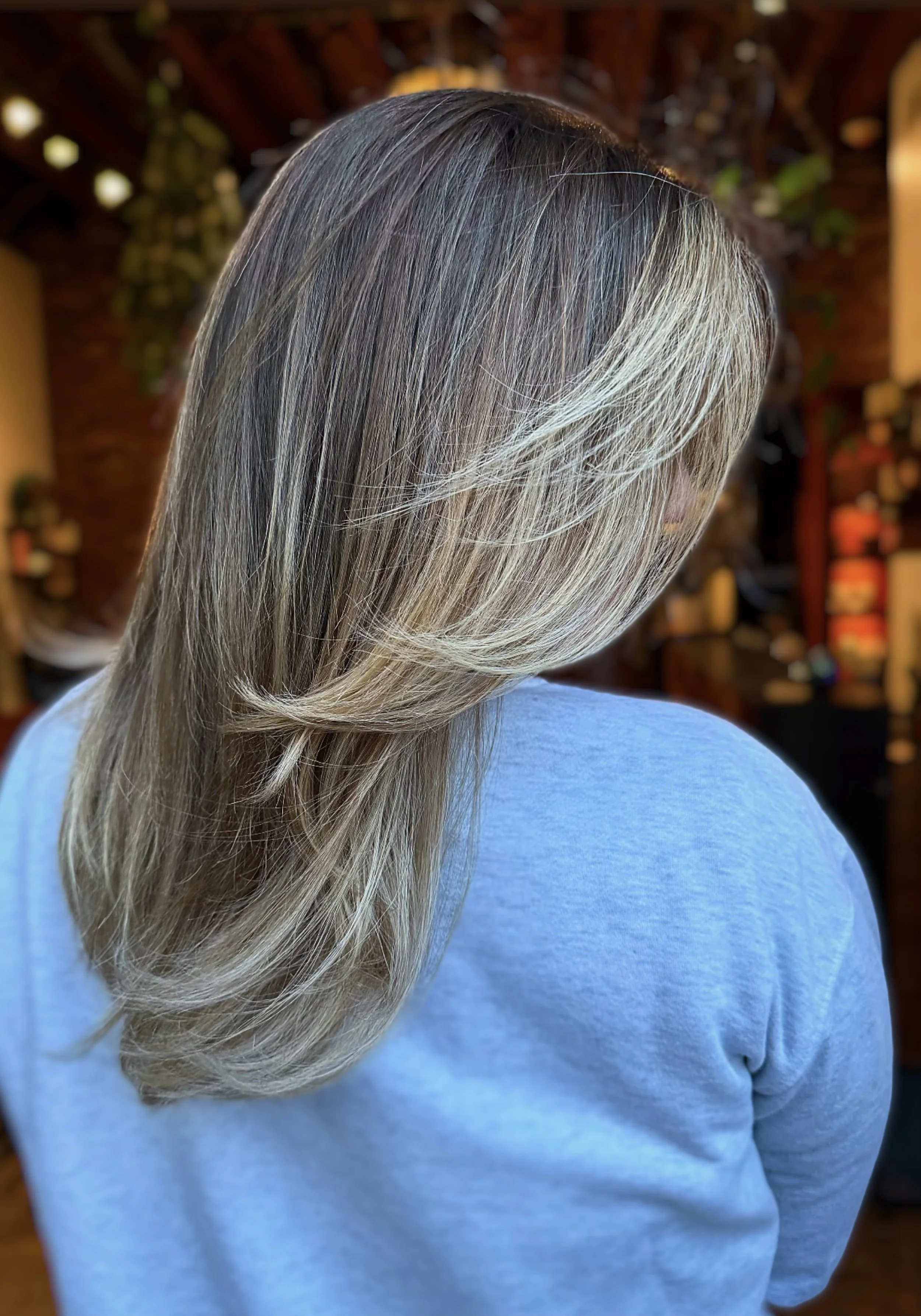 Close-up of a woman with long, layered blonde and gray hair, wearing a light blue sweater, inside a cozy indoor setting.