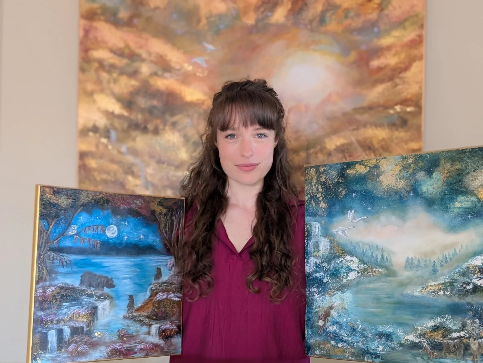 Woman with long curly brown hair wearing a maroon top, standing among three landscape paintings with nature scenes, a mountain scene, and a forest scene.