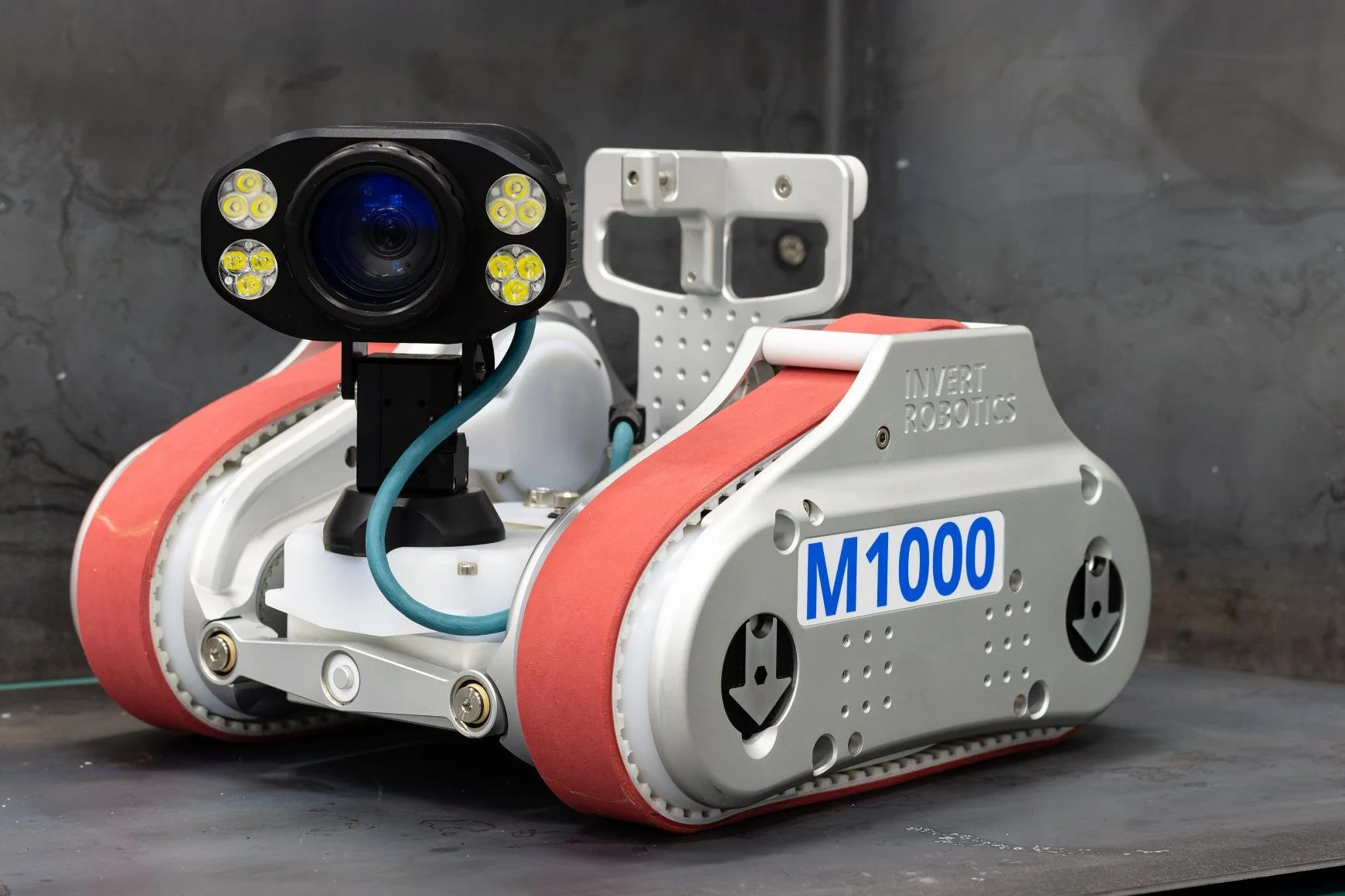 A Invert Robotics M1000 robotic vehicle with pink-colored tracks and a camera mounted on top, labeled with 'M1000' and 'INVERT ROBOTICS', positioned against a gray background.