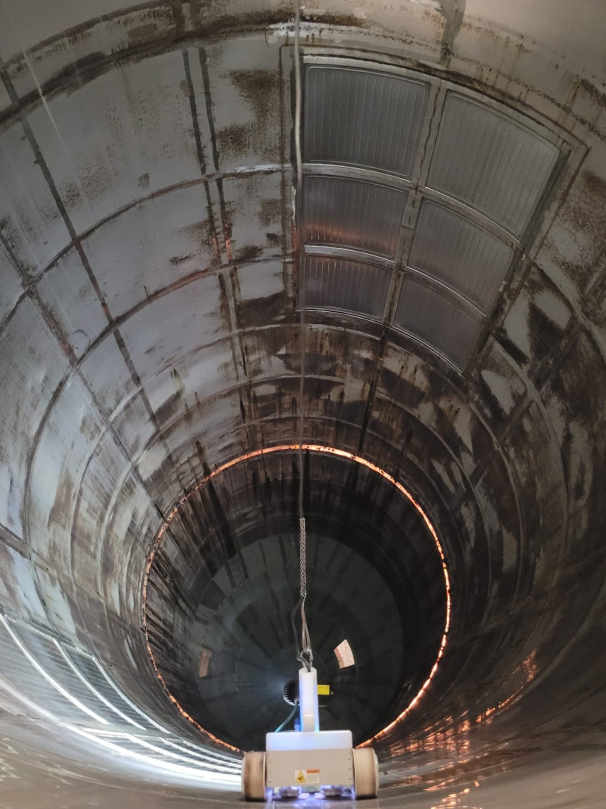 Looking inside a large industrial tunnel with a high curved ceiling, showing metallic walls with rust and dirt, and a machine at the opening with glowing lights and a white cable hanging down.