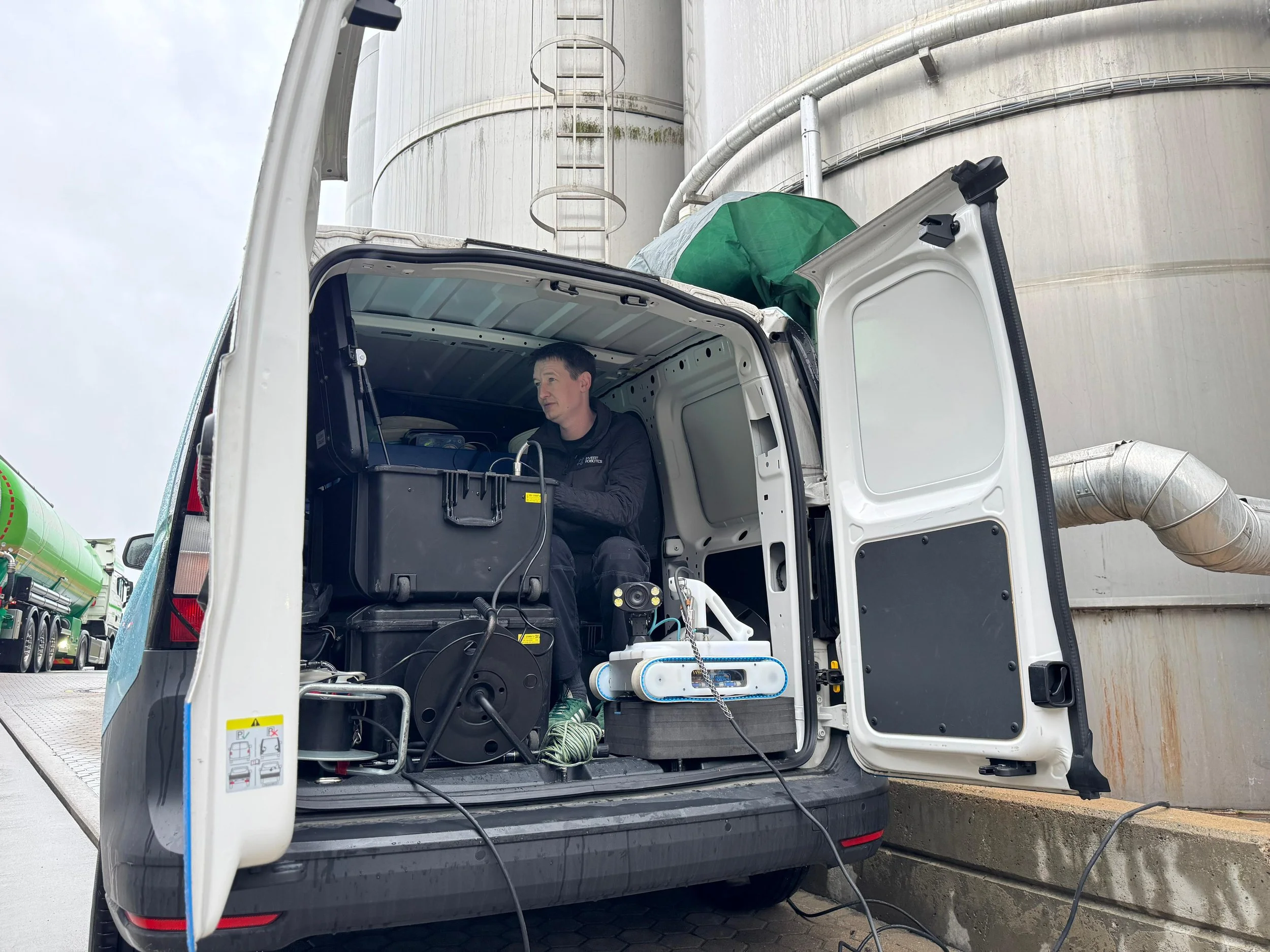 A man sitting inside a partially open white utility van, working with robotic inspection equipment. The vanis parked in an industrial area with large storage tanks and pipes visible in the background.