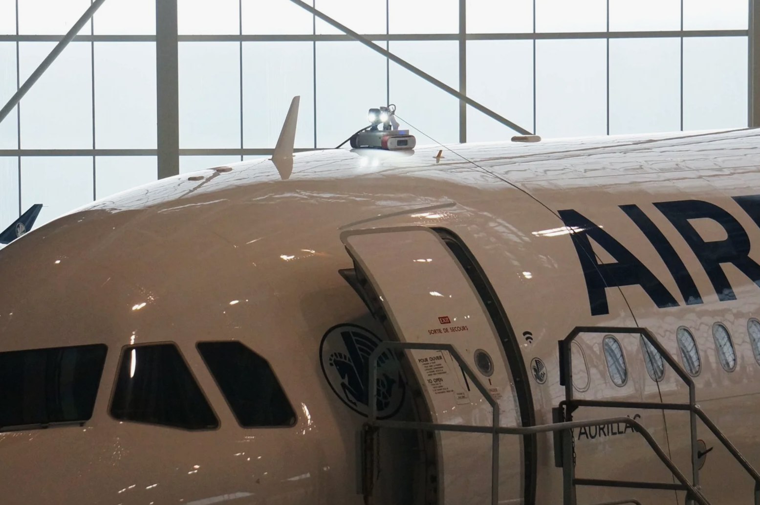 Close-up of an airplane's cockpit with a partially open door inside a spacious hangar, with large windows in the background.