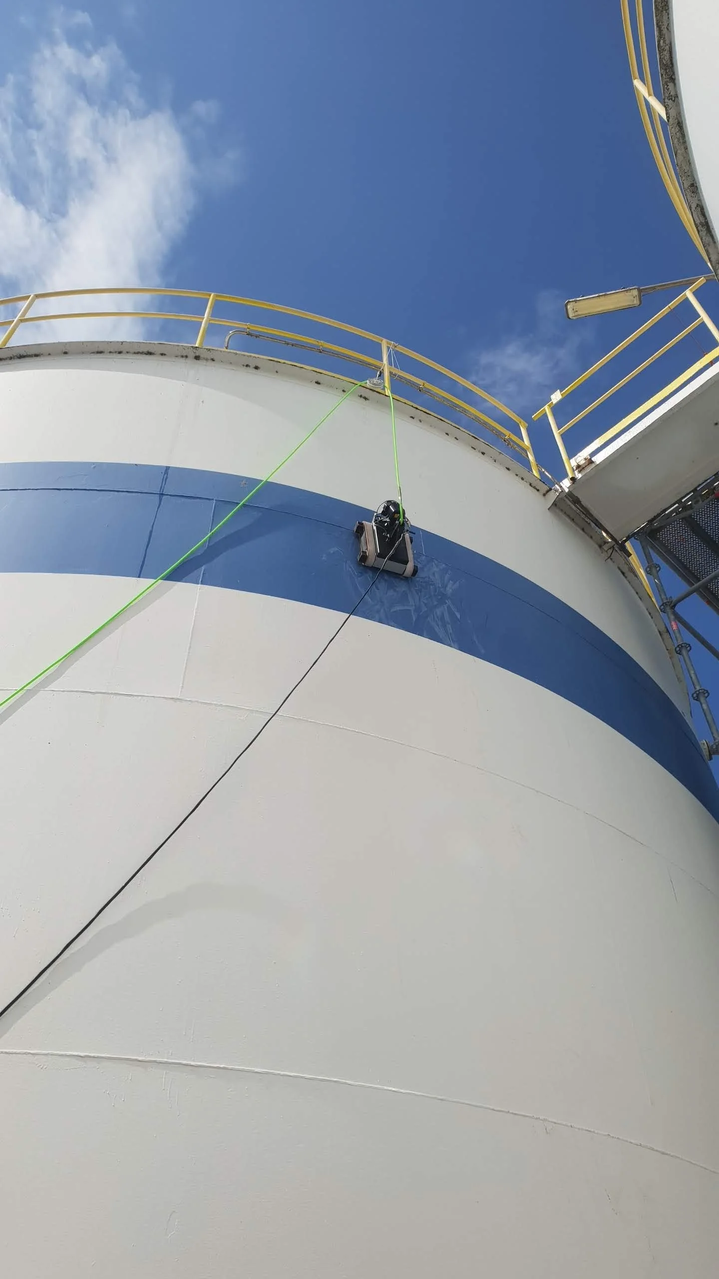 View looking up at the side of a large white industrial storage tank with a blue stripe, featuring yellow safety railing at the top, against a clear blue sky with some clouds, and a suspended cleaning or maintenance device attached to the tank's side