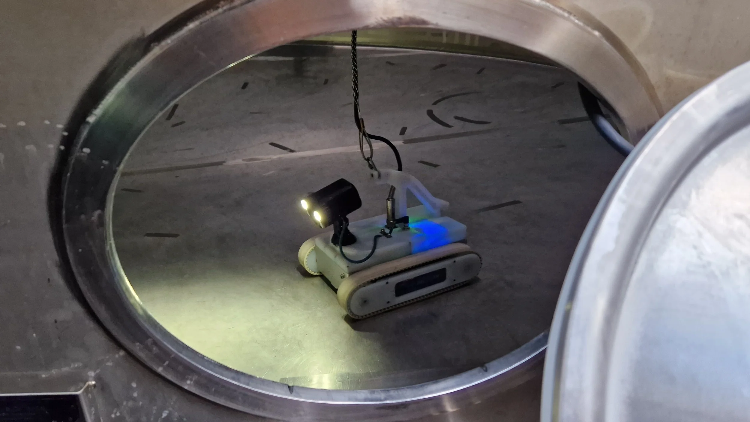 Inside a laboratory or industrial setting, a small robotic device with LED lights is placed on the floor, connected to a wire hanging from above.