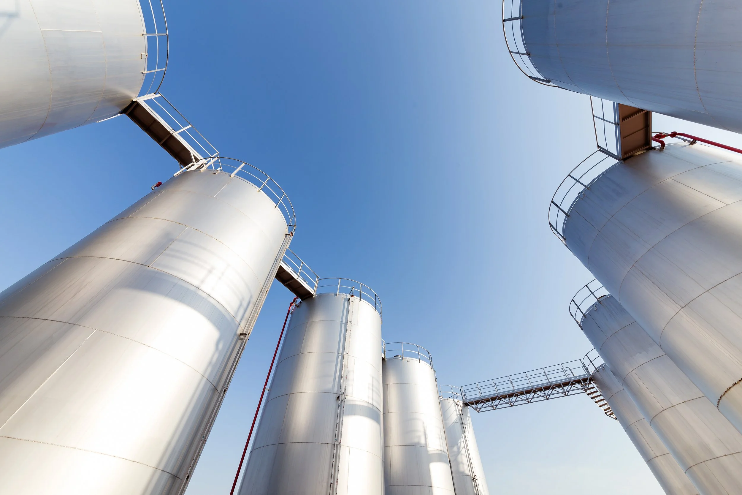 Looking up at four large, silver industrial storage tanks with stairs and railings against a clear blue sky.