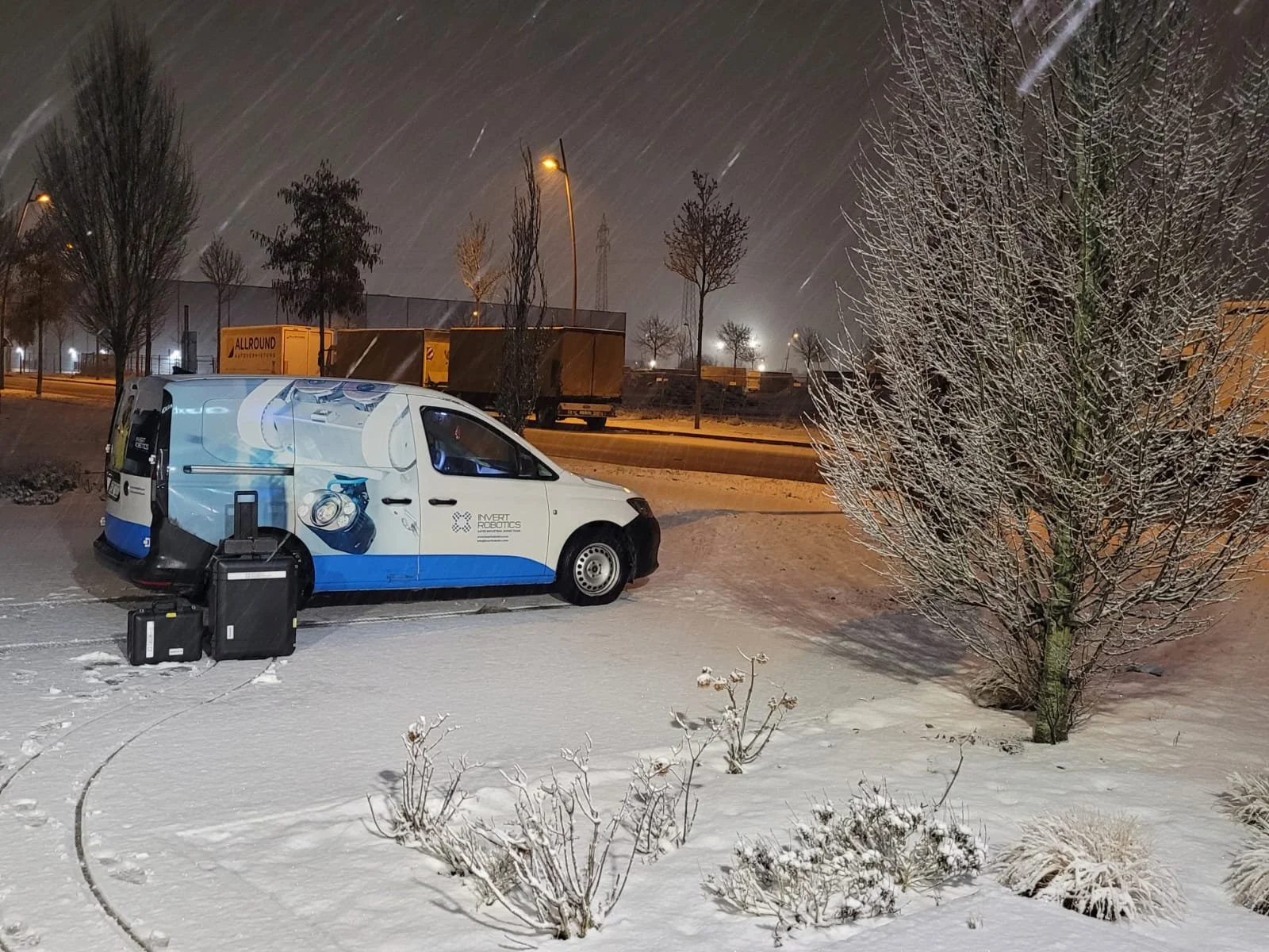 A white research vehicle with logos and graphics parked on snow-covered ground near some small bushes and a leafless tree. Two black suitcases are on the snow next to the vehicle. In the background, there are trees, streetlights, and trucks on a road