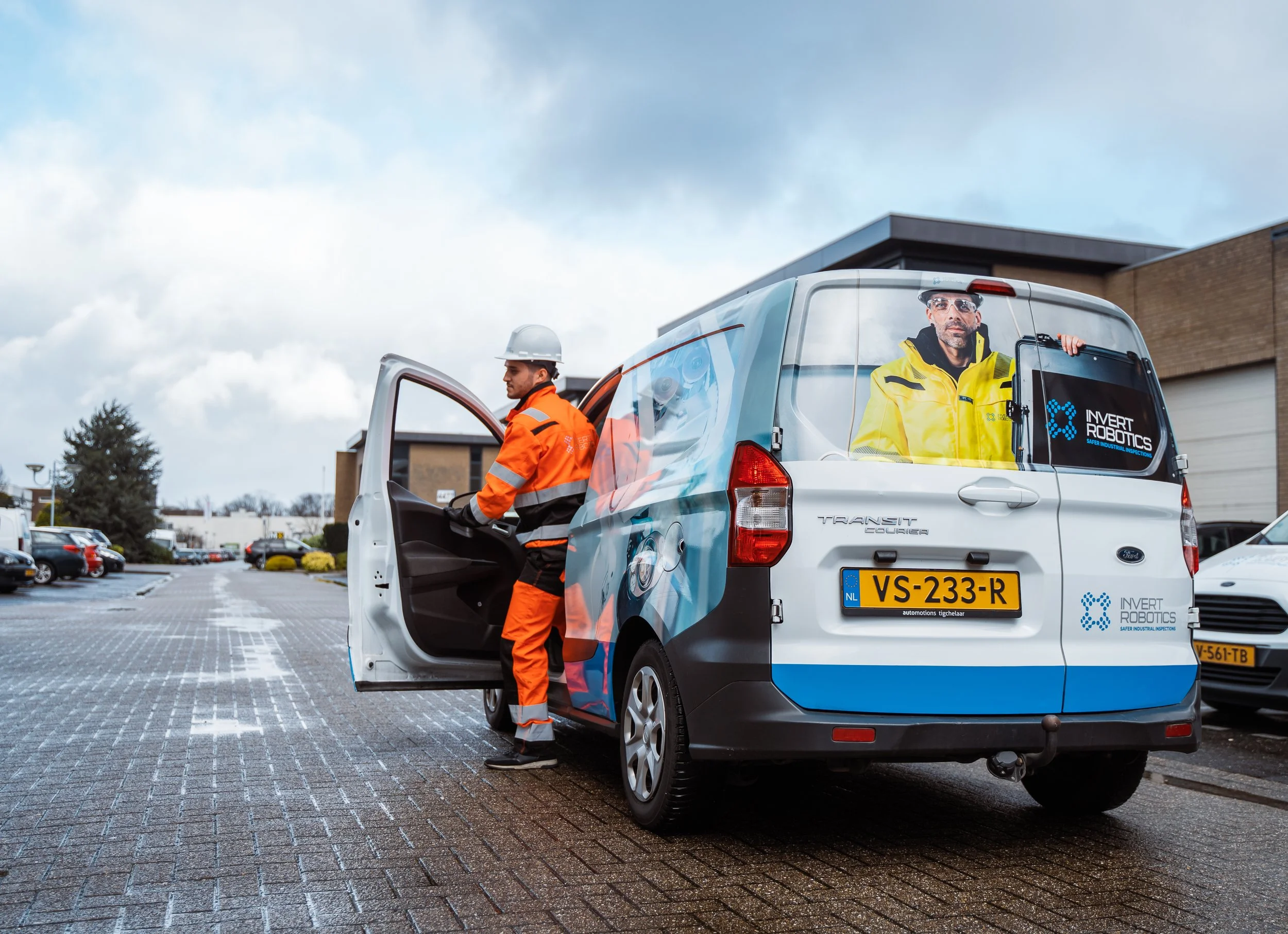 Two workers in safety gear, one inside a van and the other standing outside, next to a branded autonomous vehicle labeled 'Invert Robotics' with a cityscape in the background.