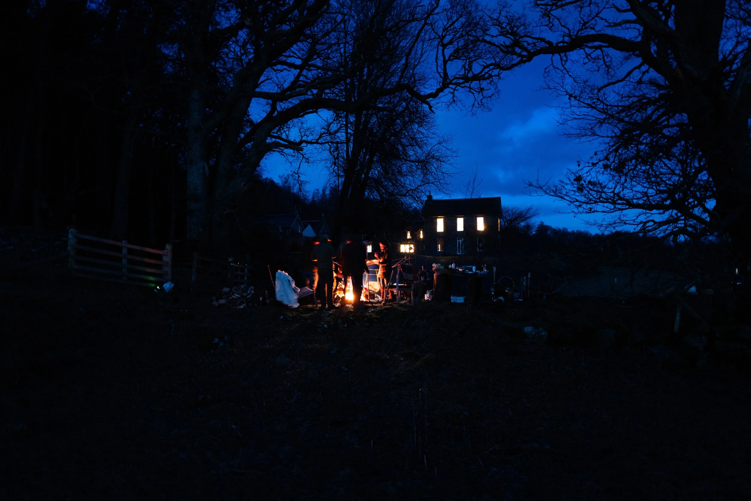 A nighttime outdoor scene with a group of people gathered around a campfire under large, bare trees near a house with illuminated windows.