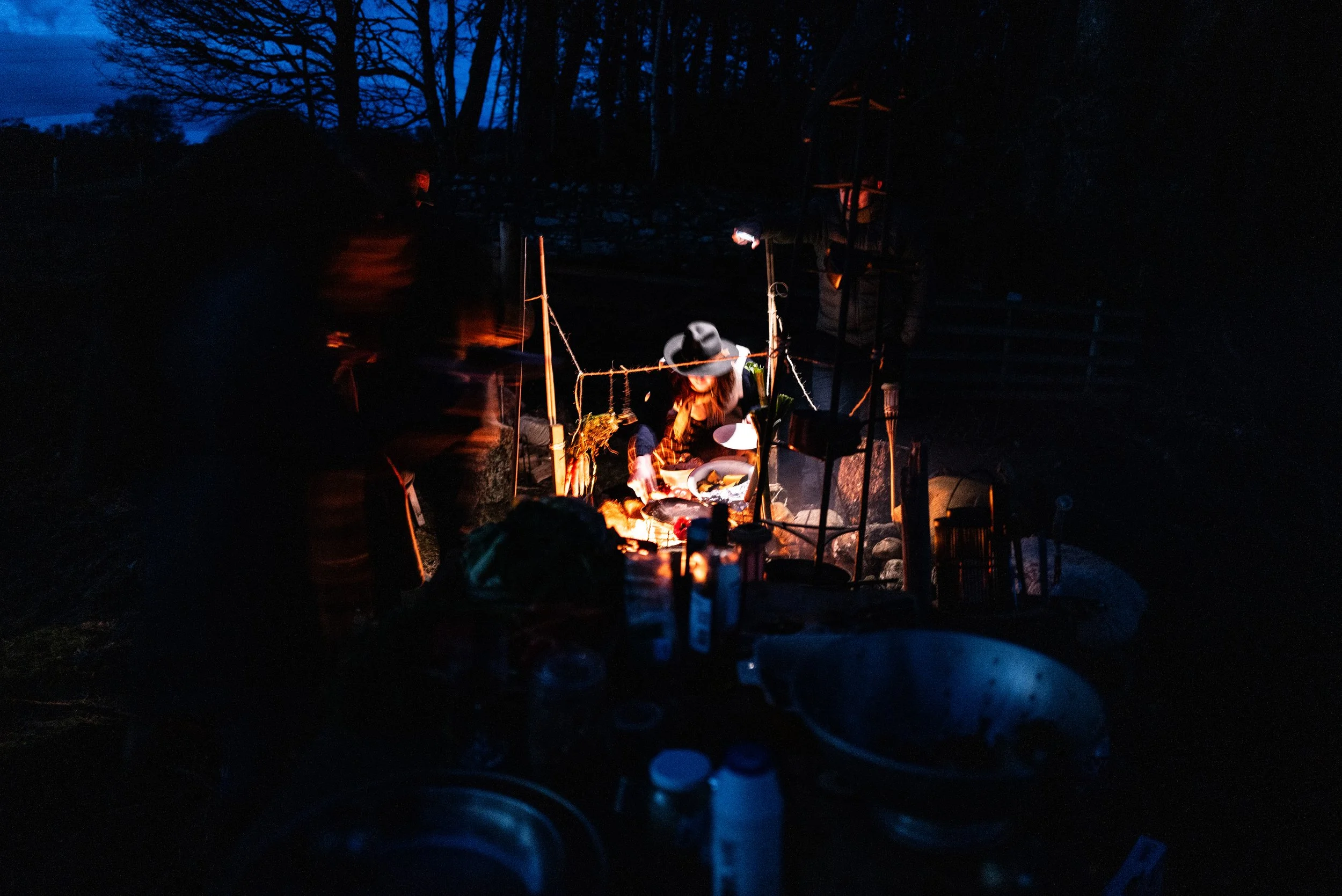 Nighttime outdoor scene with a person cooking or serving food under a canopy light, surrounded by trees and outdoor equipment.
