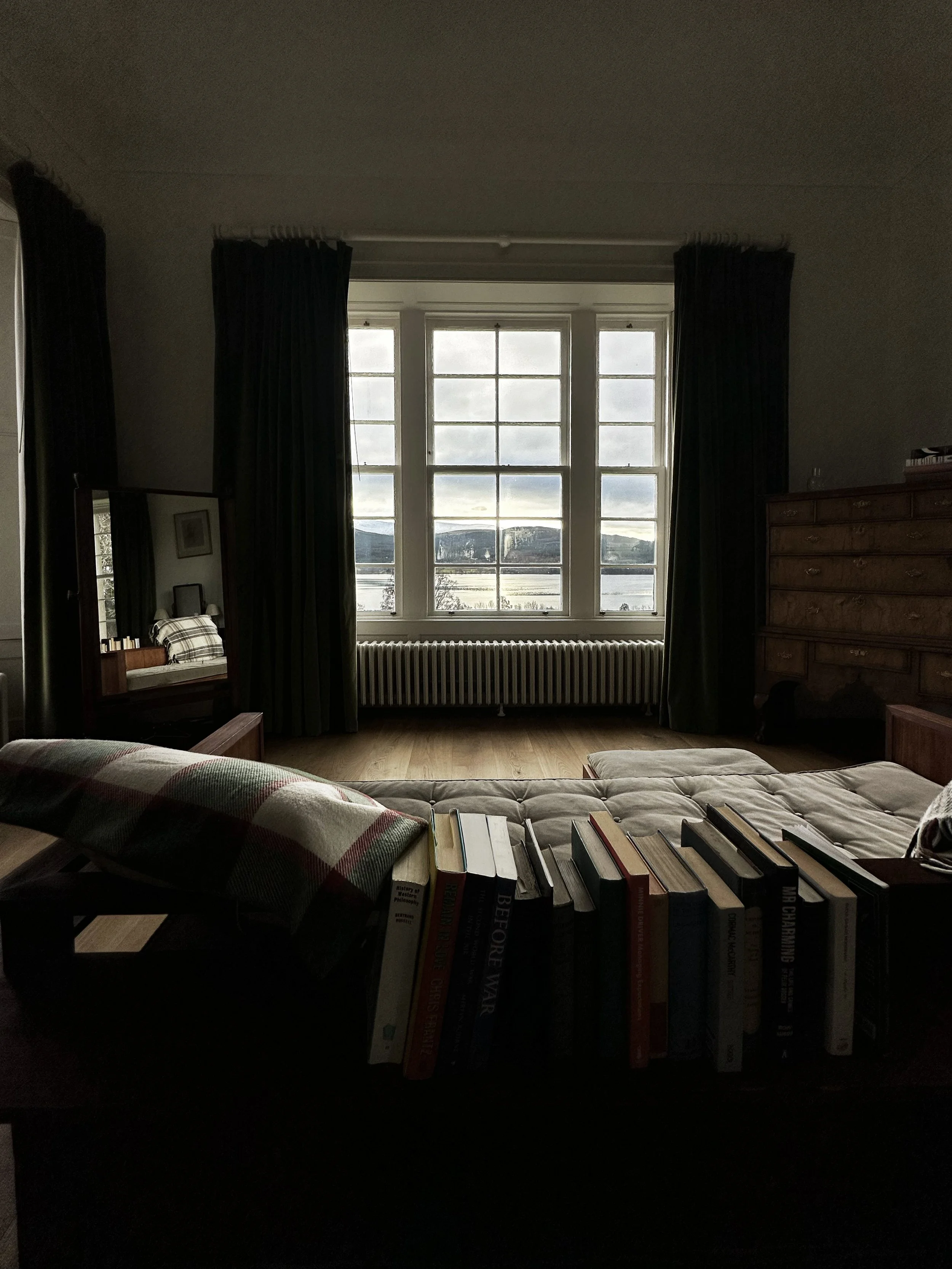 Interior room with large window showing a view of water and hills, dark curtains, wooden furniture, a bed with a plaid blanket, a bookshelf with books, and a mirror reflecting part of the bed.