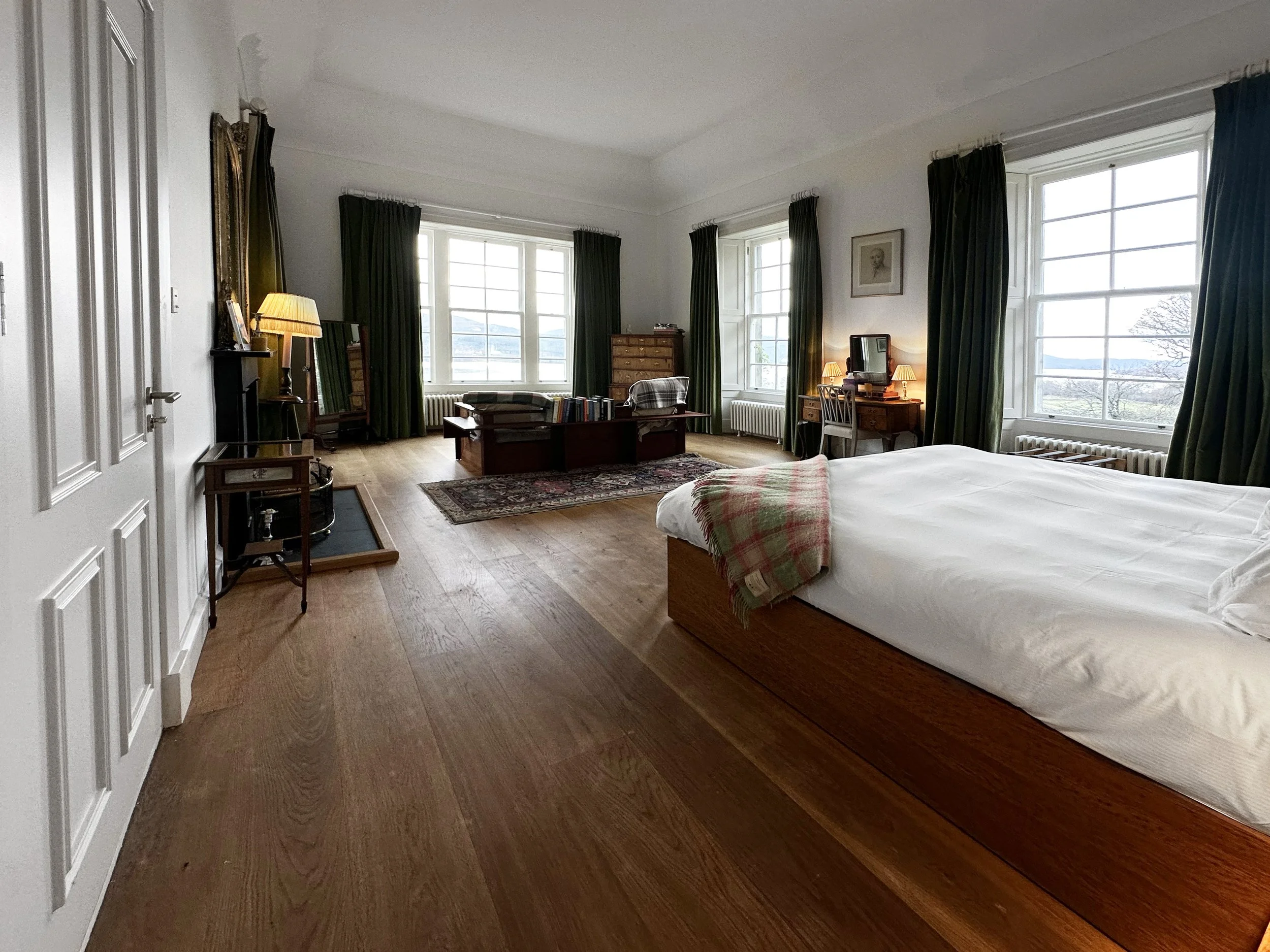 A spacious bedroom with large windows, a wooden floor, a bed with a plaid blanket, a desk with a mirror and a chair, and a small table with a lamp.
