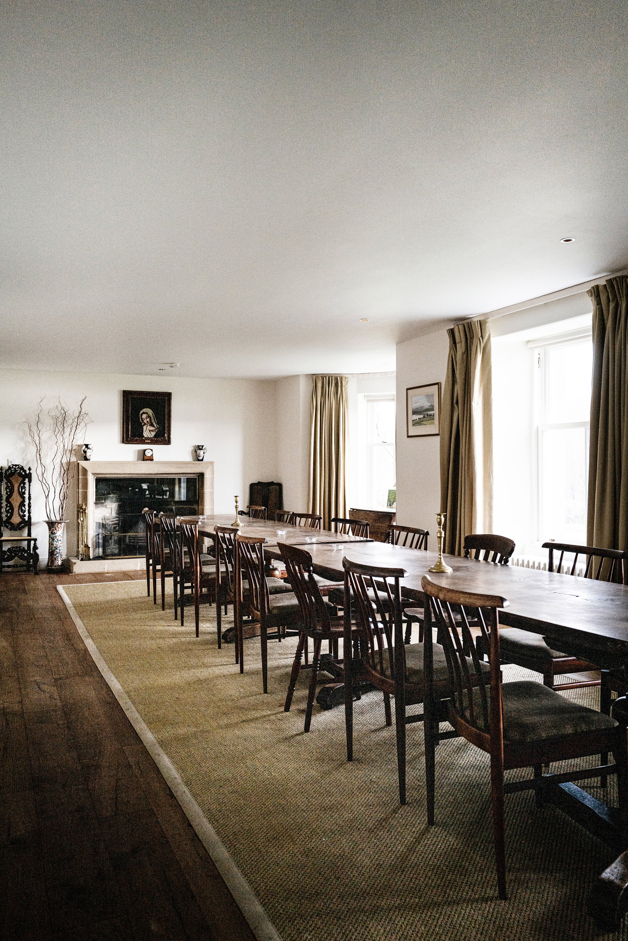A dining room with a long wooden table, surrounded by matching wooden chairs. There are large windows with cream curtains, and framed pictures on the walls. The room has a fireplace with artwork above it, and decorative vases and branches on the mantle. The floor is dark wood with a light-colored rug underneath the table.