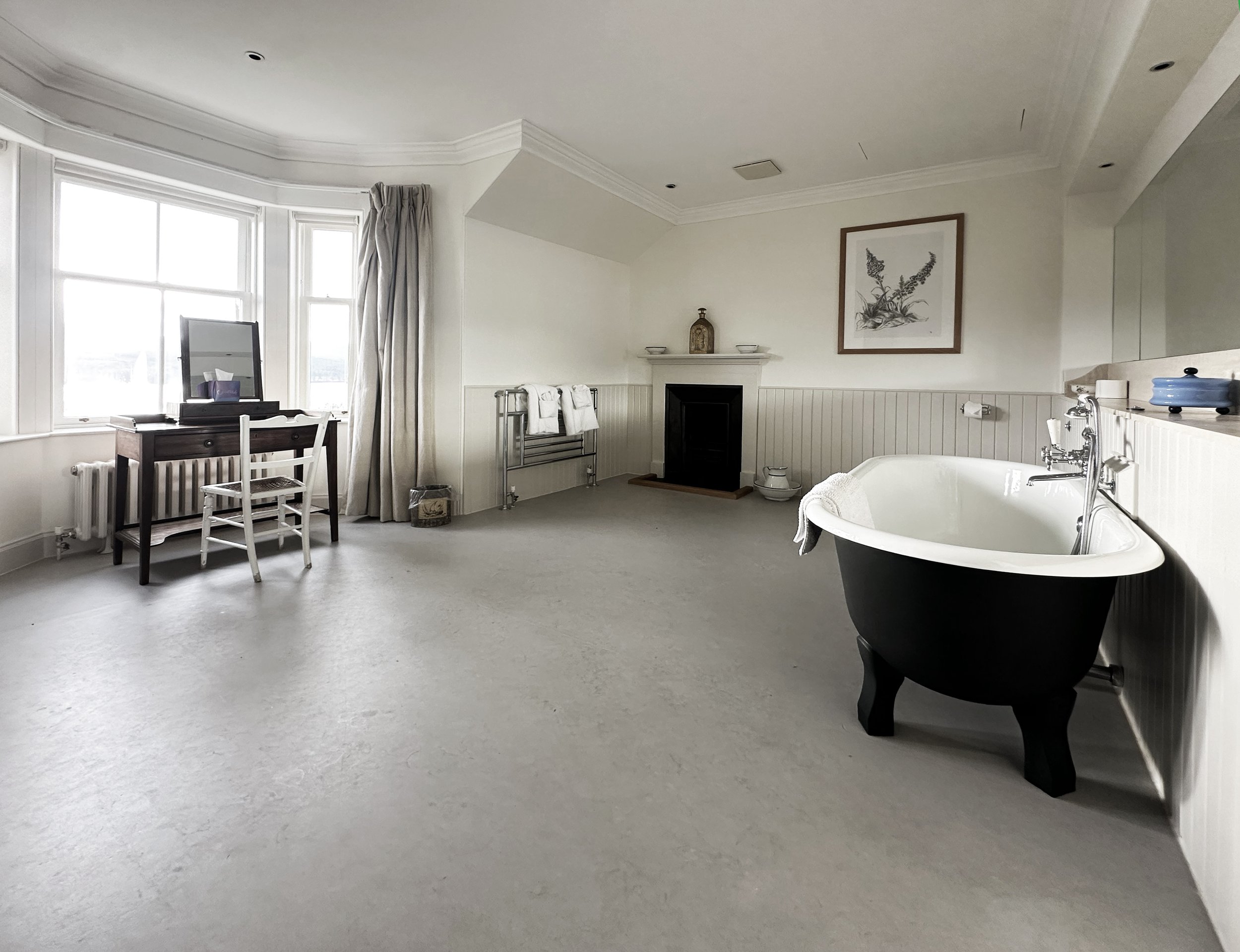 A bathroom with a white bathtub, towels, a small table with a mirror, window with curtains, and wall art of a plant.
