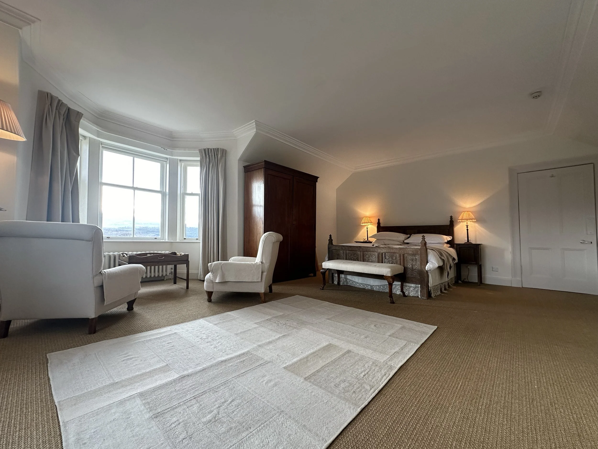 A spacious bedroom with a large window, beige carpet, white walls, a bed with patterned bedding, nightstands with lamps, a wooden wardrobe, white armchairs, and a white rug on the floor.