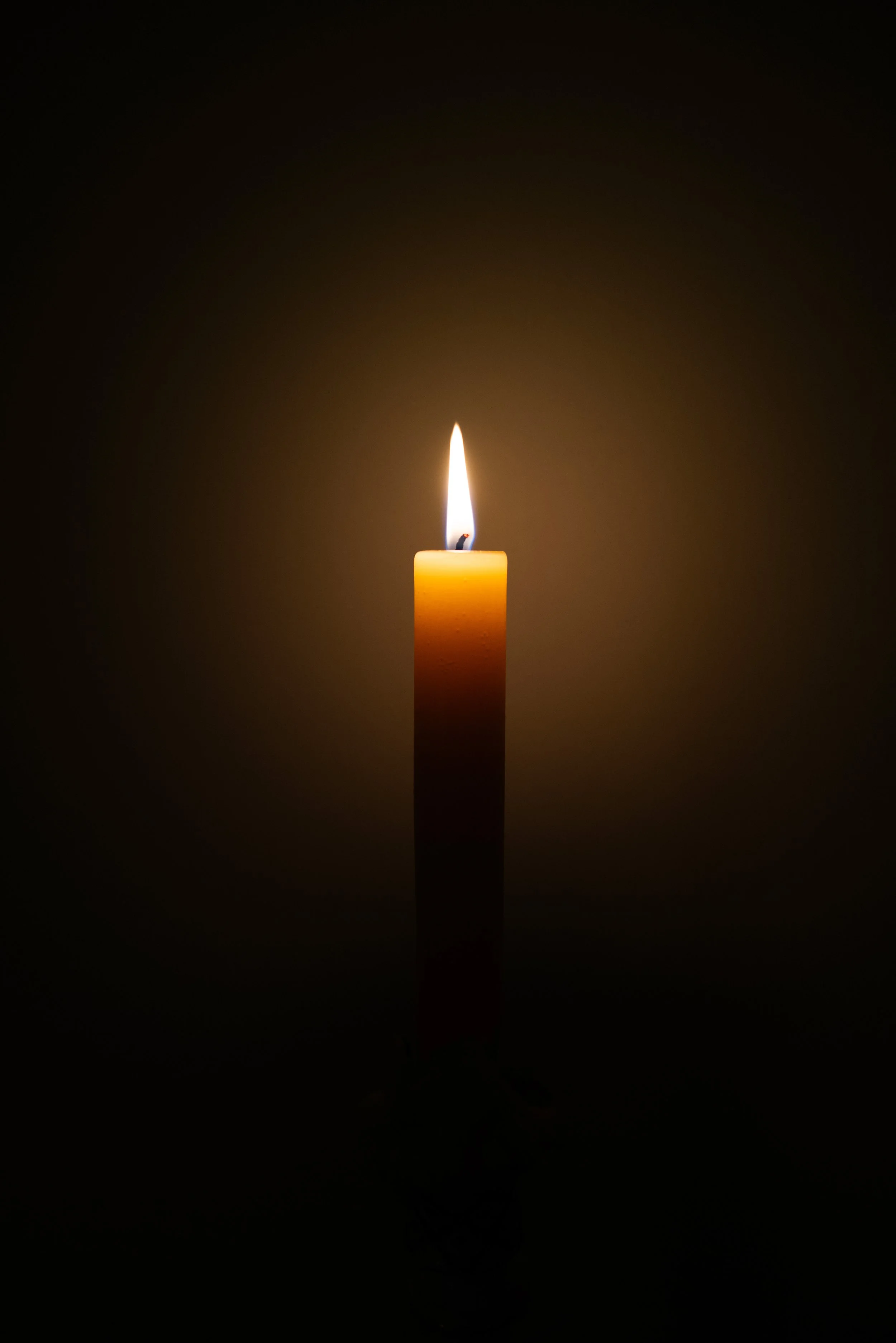 A lit yellow candle against a black background with the flame glowing and illuminating the surrounding area.