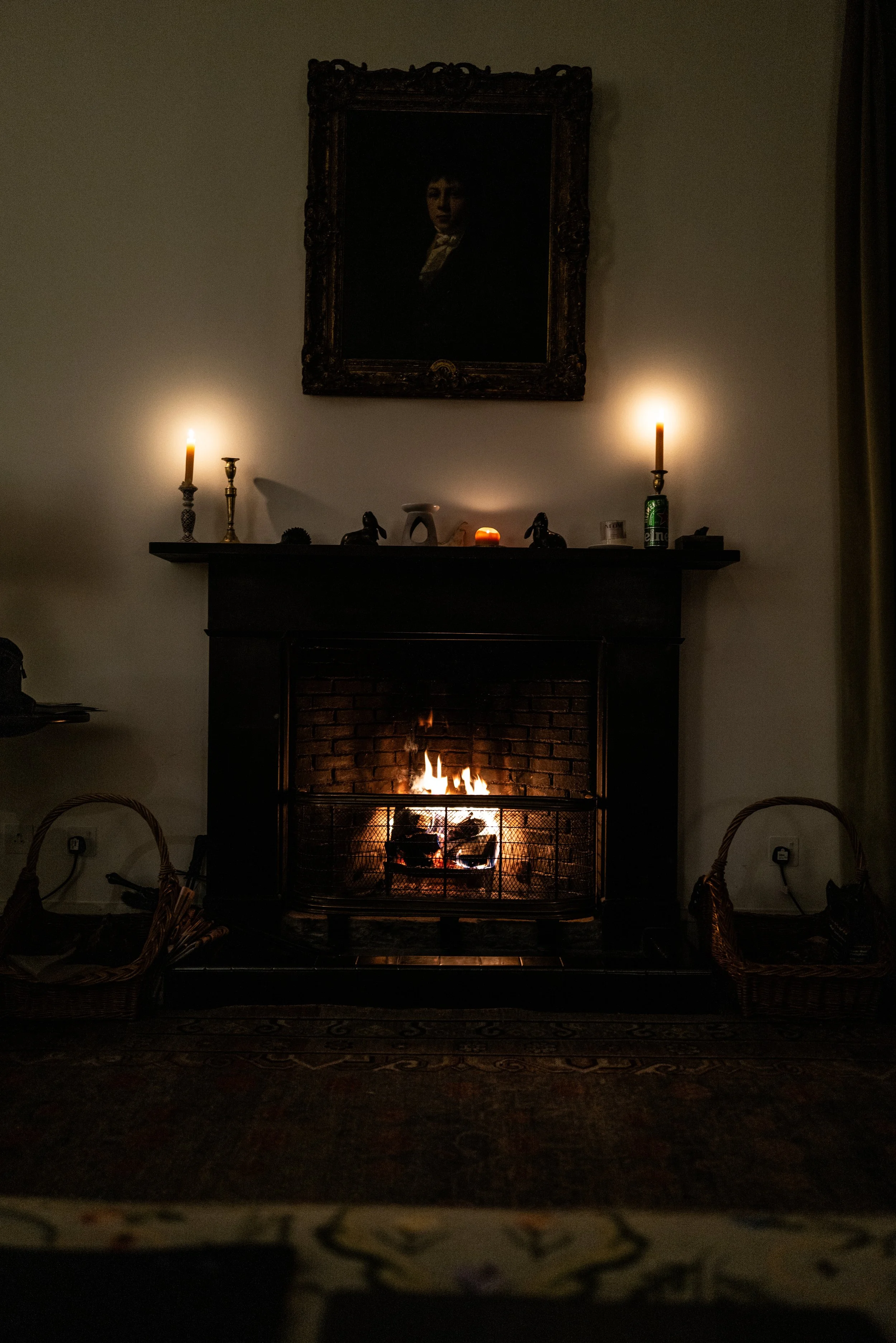 Living room with a brick fireplace, lit with a fire, a framed portrait above the mantle, and candles on the mantle. There are wicker chairs and a patterned rug.