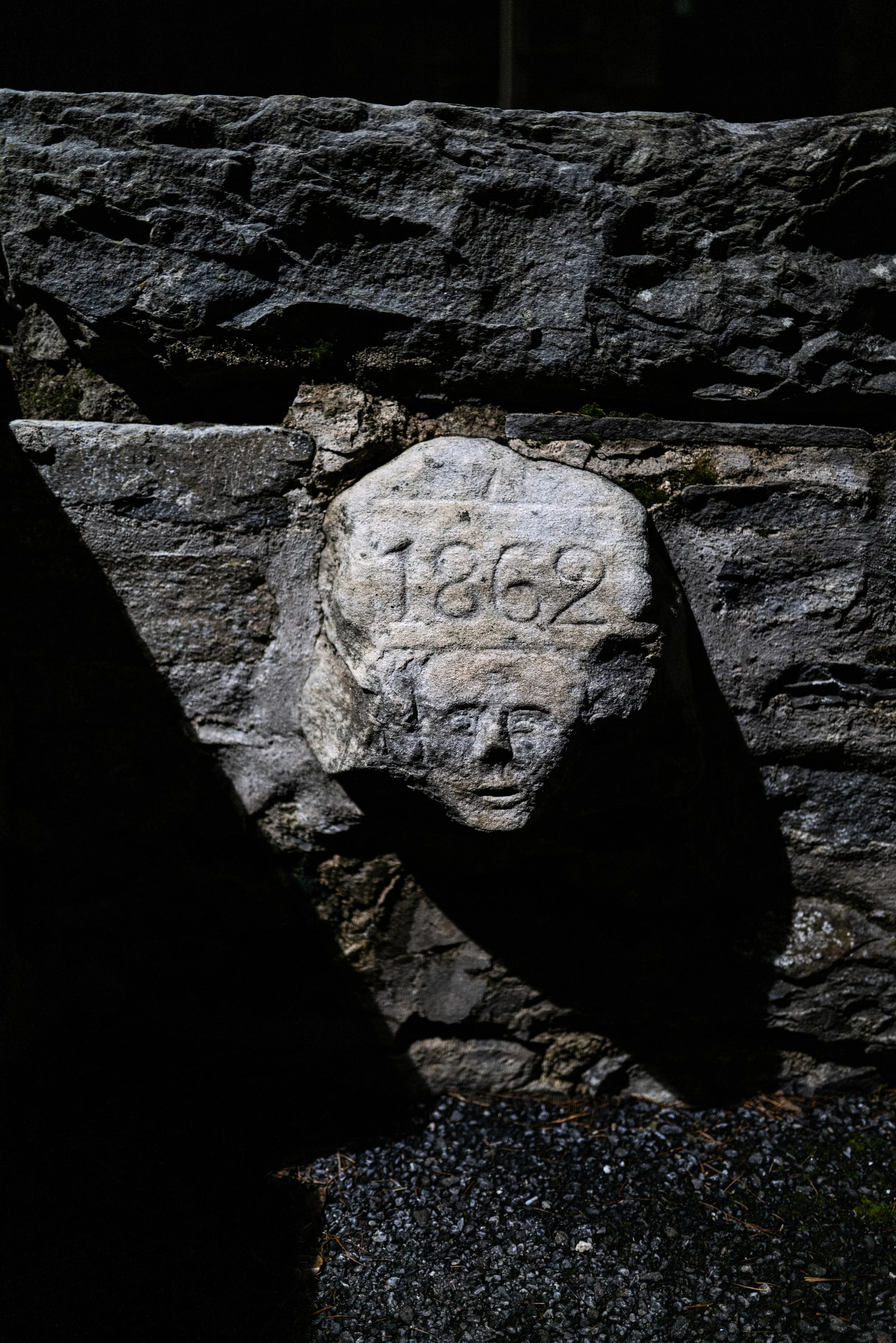 Close-up of a textured stone wall with a carved face and the year "1862" engraved on it, illuminated by sunlight.