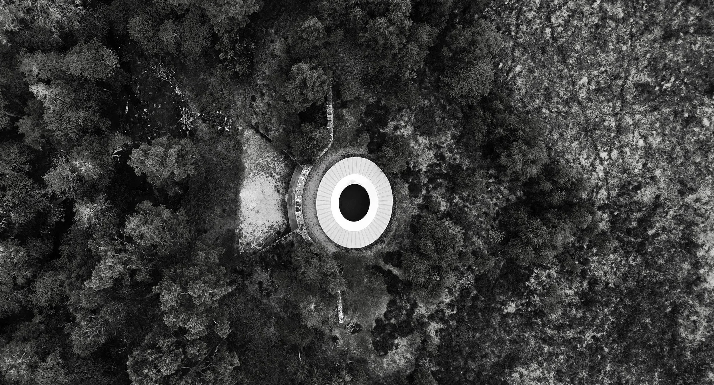 An aerial black and white photograph of a circular building with a large oval opening in the center, surrounded by a dense forest.