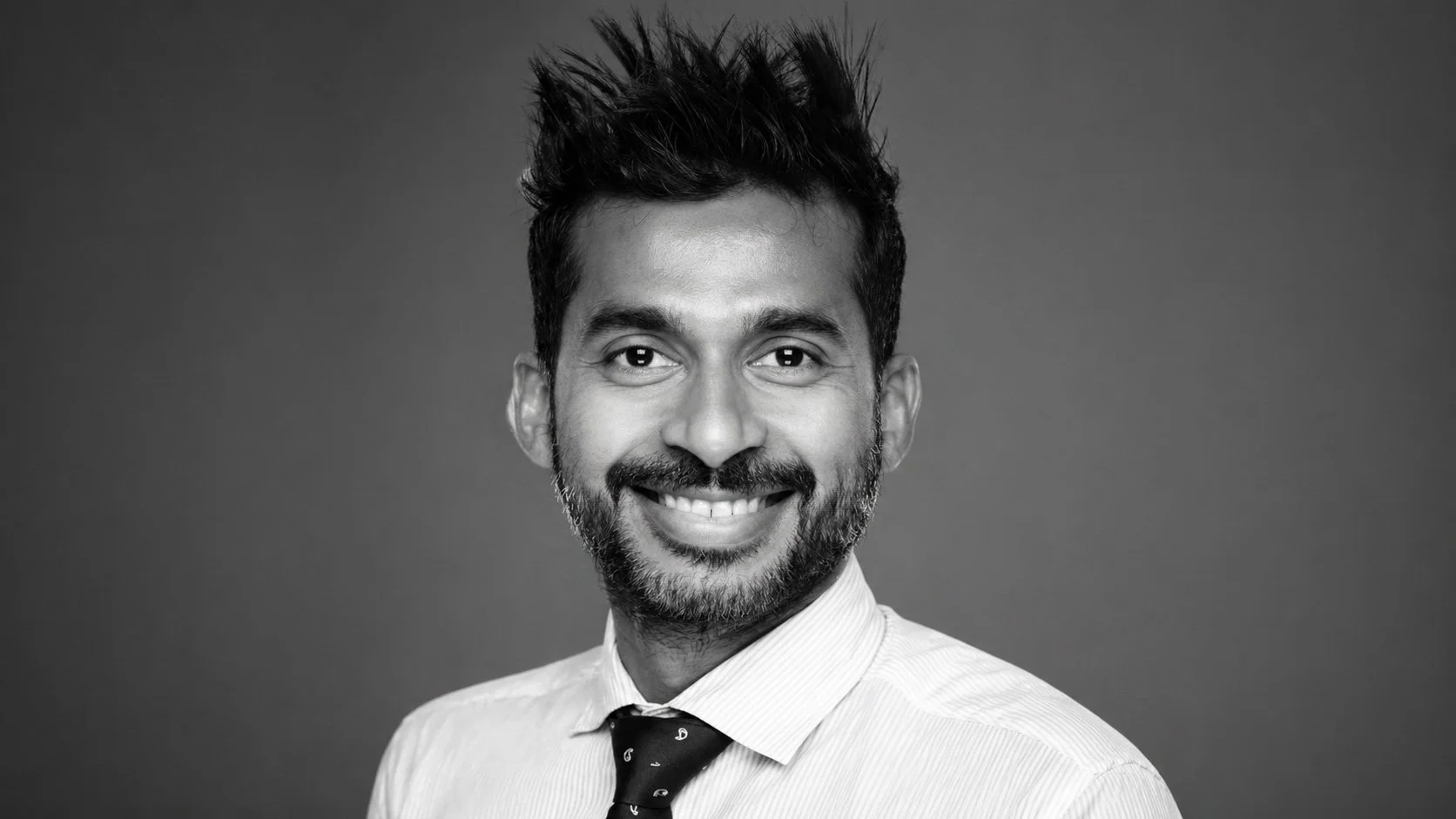 Black and white headshot of a man with a smile, groomed beard, spiky hair, wearing a collared shirt and a tie, against a plain background.