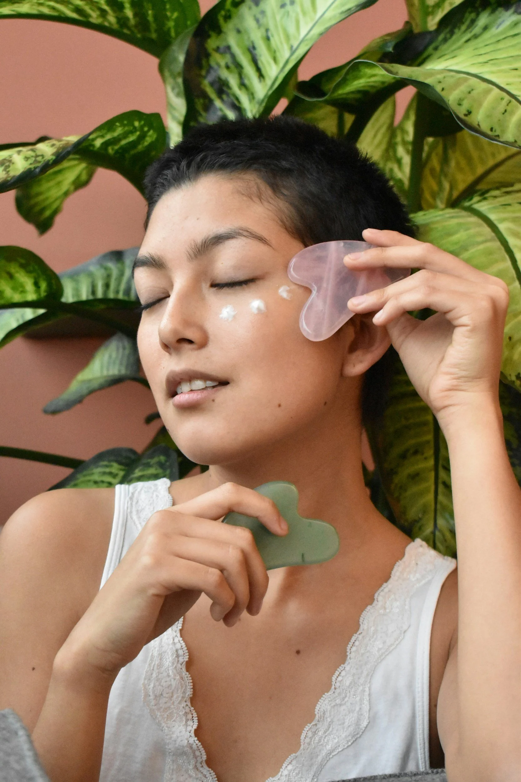 A woman with short black hair and a white lace-trimmed tank top is gently pressing a gua sha facial massage tool against her cheek, with her eyes closed and a peaceful expression.