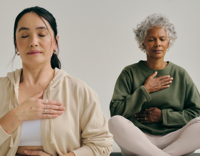 Two women seated cross-legged on a yoga mat with hands on heart and belly during a Soulibrium breathwork practice