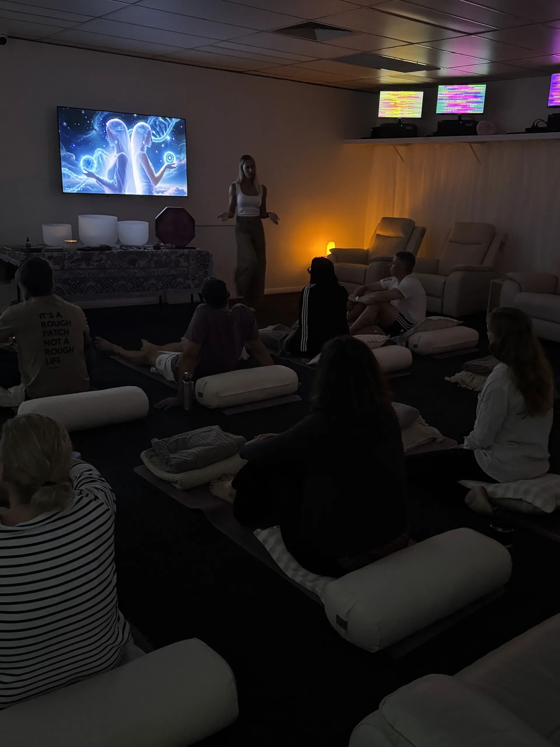 A woman stands in a dimly lit room giving a presentation to a small audience seated on floor cushions and ottomans. A large screen displays an animated image of two women with long hair, holding orbs, surrounded by a blue and purple cosmic background. The room has multiple TVs or monitors mounted on the wall, some displaying colorful, abstract patterns.
