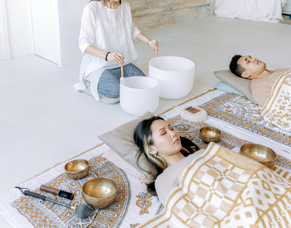 Two people lying down on mats with blankets, eyes closed, while a third person kneels playing singing bowls with a mallet, in a peaceful, indoor setting.