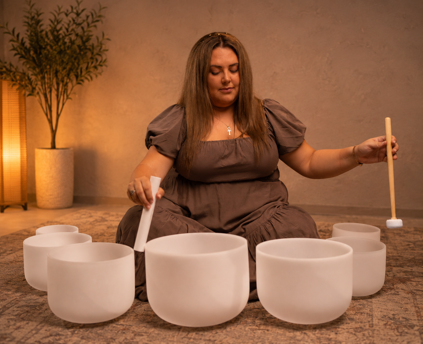 A woman sitting on the floor playing a set of white crystal singing bowls with mallets, in a room with warm lighting and a potted plant in the background.