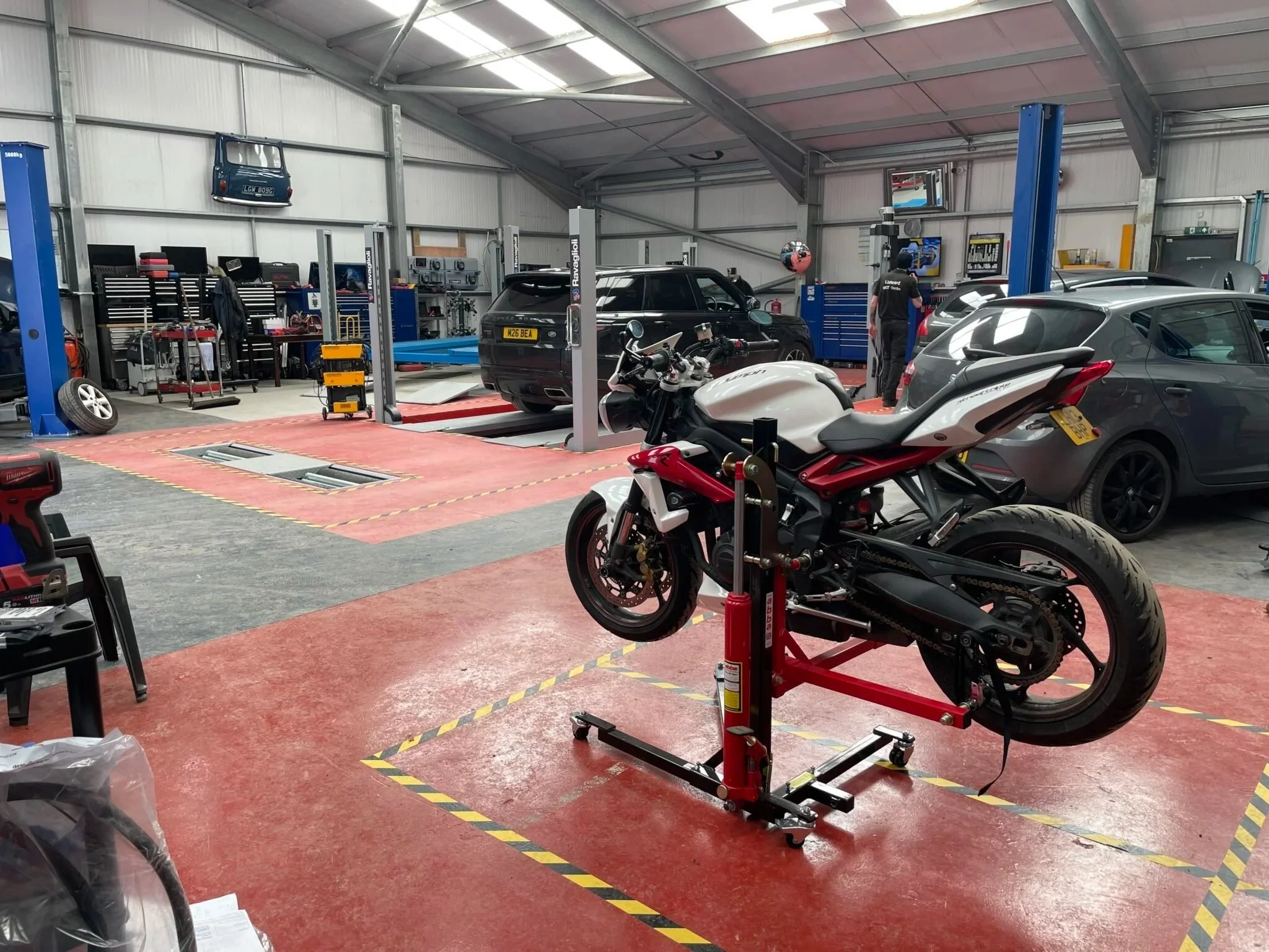 An automotive workshop with cars parked inside and a motorcycle on a stand in the foreground. Tools and equipment are organized along the back wall, and a worker is attending to a car on a lift. The space has a high ceiling with skylights, and the floor is marked with yellow and black caution tape.