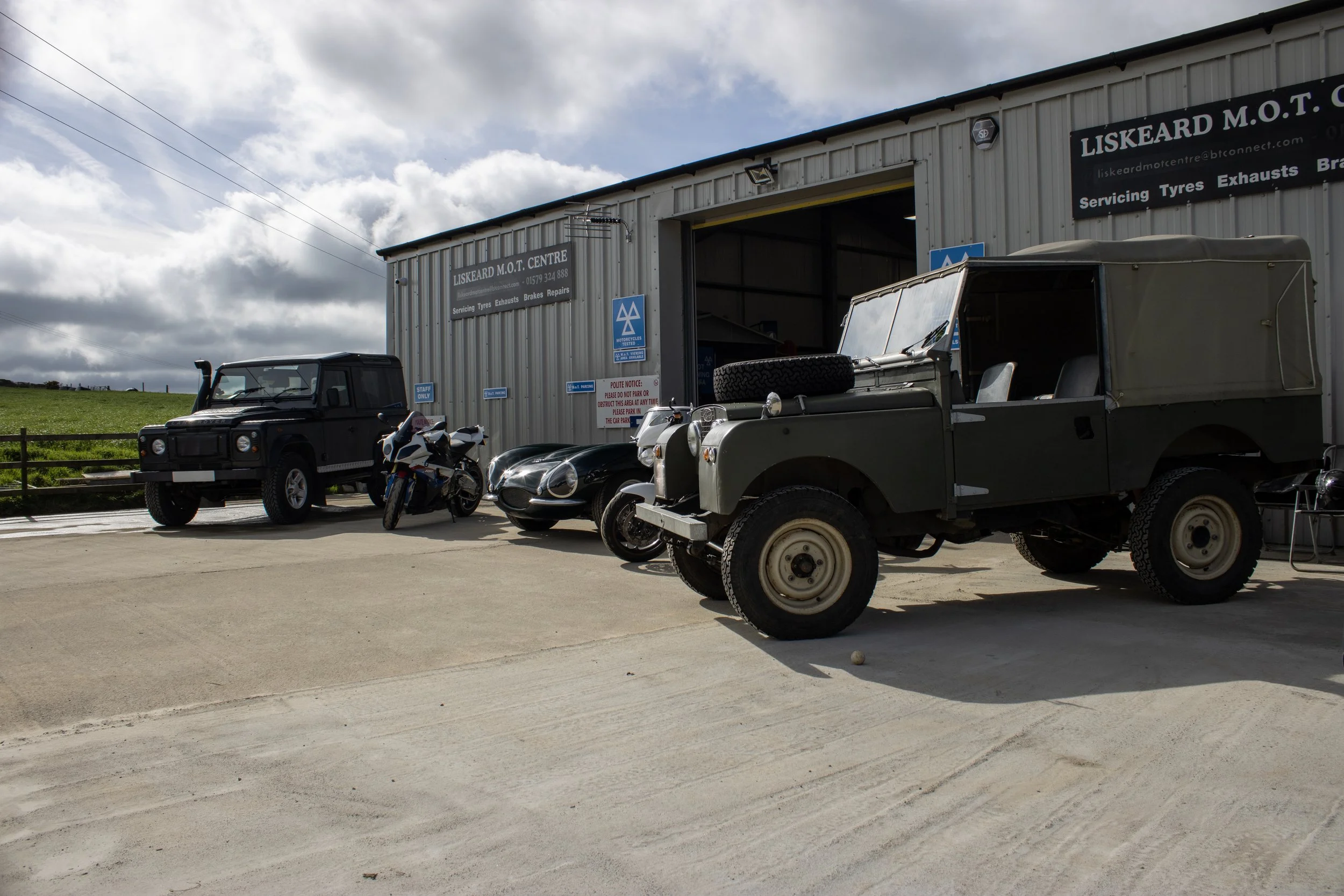 Outside view of a garage with vintage vehicles and motorcycles parked nearby under a partly cloudy sky.