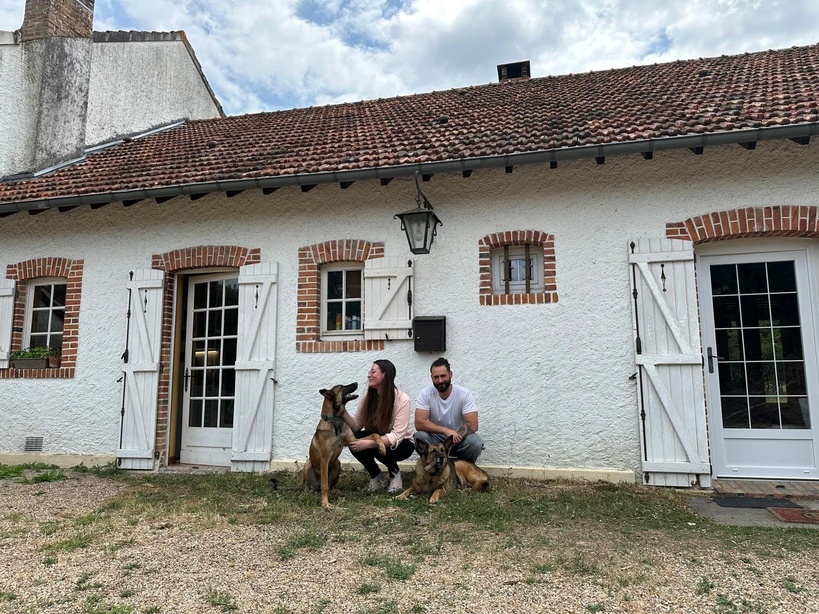 Une maison blanche avec des fenêtres à volets en bois blanc, un toit en tuiles rouges, et deux personnes assises devant avec deux chiens.