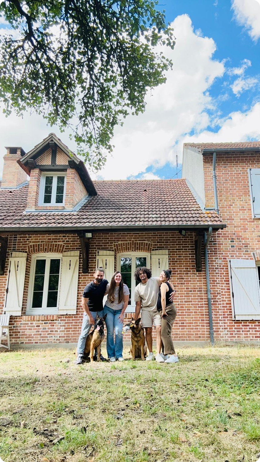 Groupe de quatre personnes souriantes posant avec deux chiens devant une maison en briques avec un jardin, sous un ciel bleu avec des nuages.