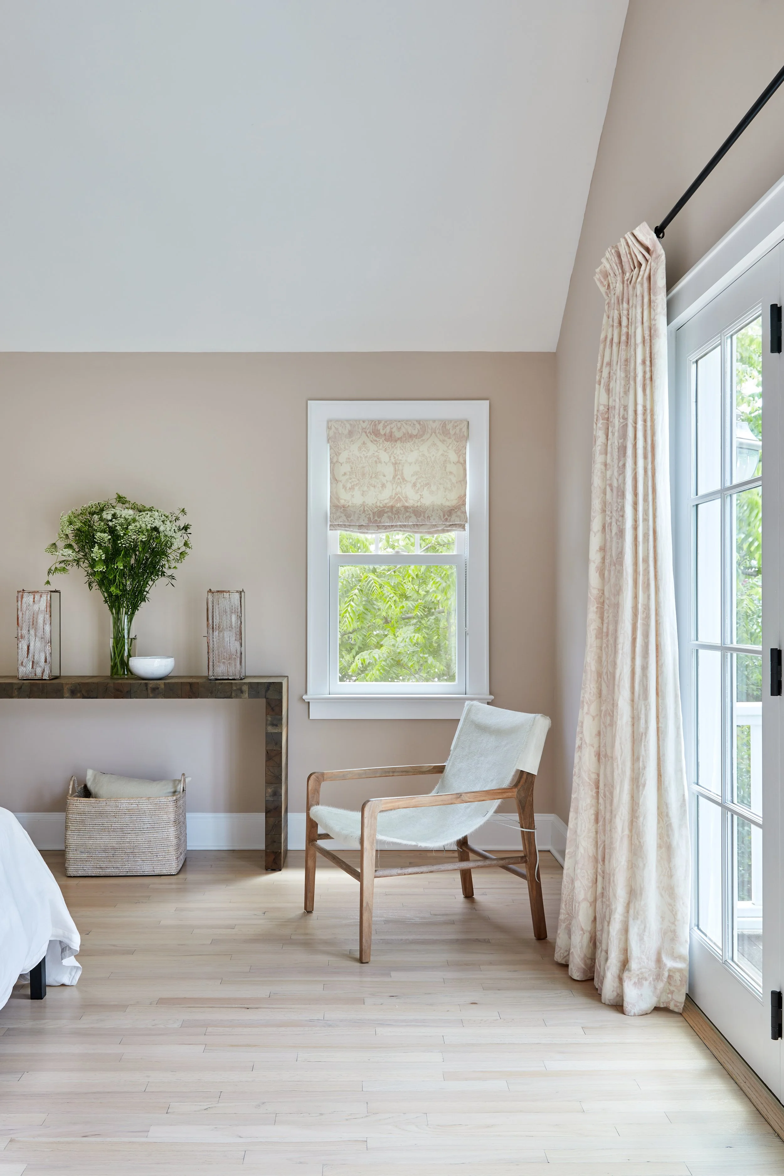 Considered bedroom corner with natural wood lounge chair, patinated console table, layered window treatments, and organic accessories by Ginger Lemon Indigo